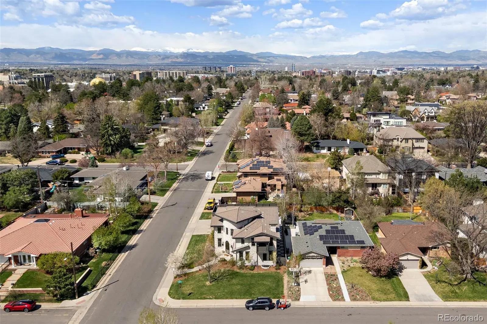 This aerial view showcases a well-established residential neighborhood with a mix of architectural styles. The homes feature manicured lawns, mature trees, and some have solar panels. In the distance, a city skyline is visible against a backdrop of mountains, creating a picturesque setting.