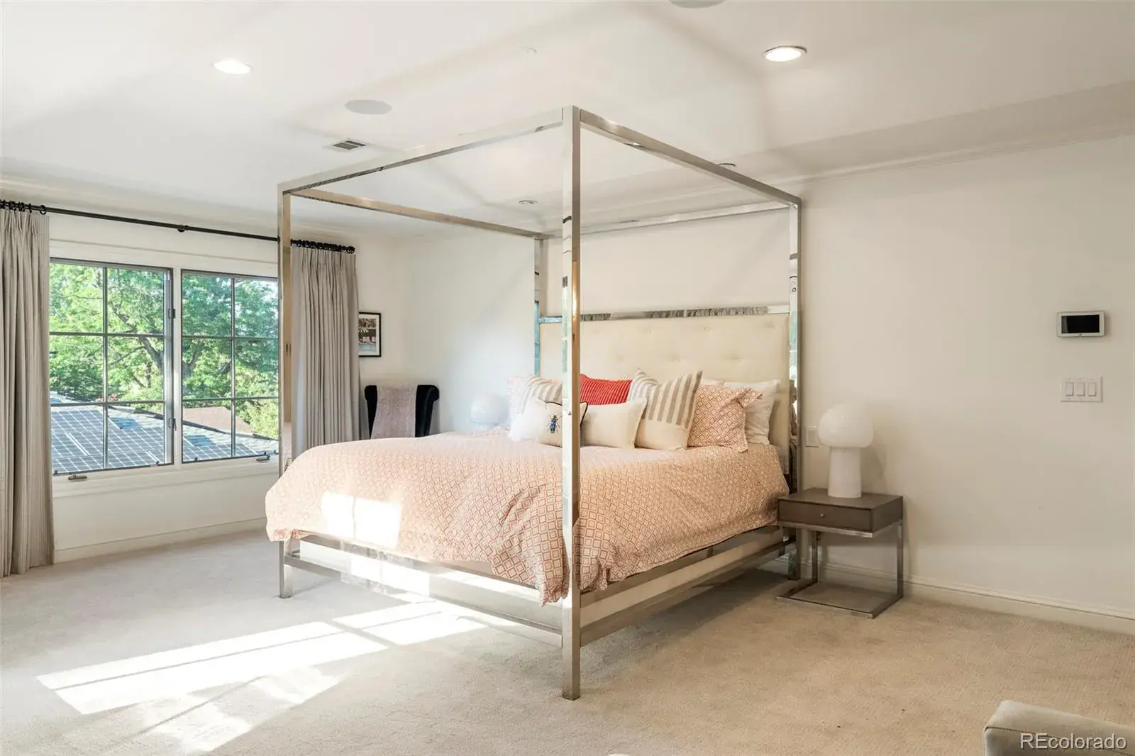 This is a bright and airy primary bedroom featuring a four-poster bed with a light-colored headboard and patterned bedding. Natural light floods the room through a large window, complemented by neutral-toned walls and carpet. A bedside table with a lamp adds a touch of elegance to the space.