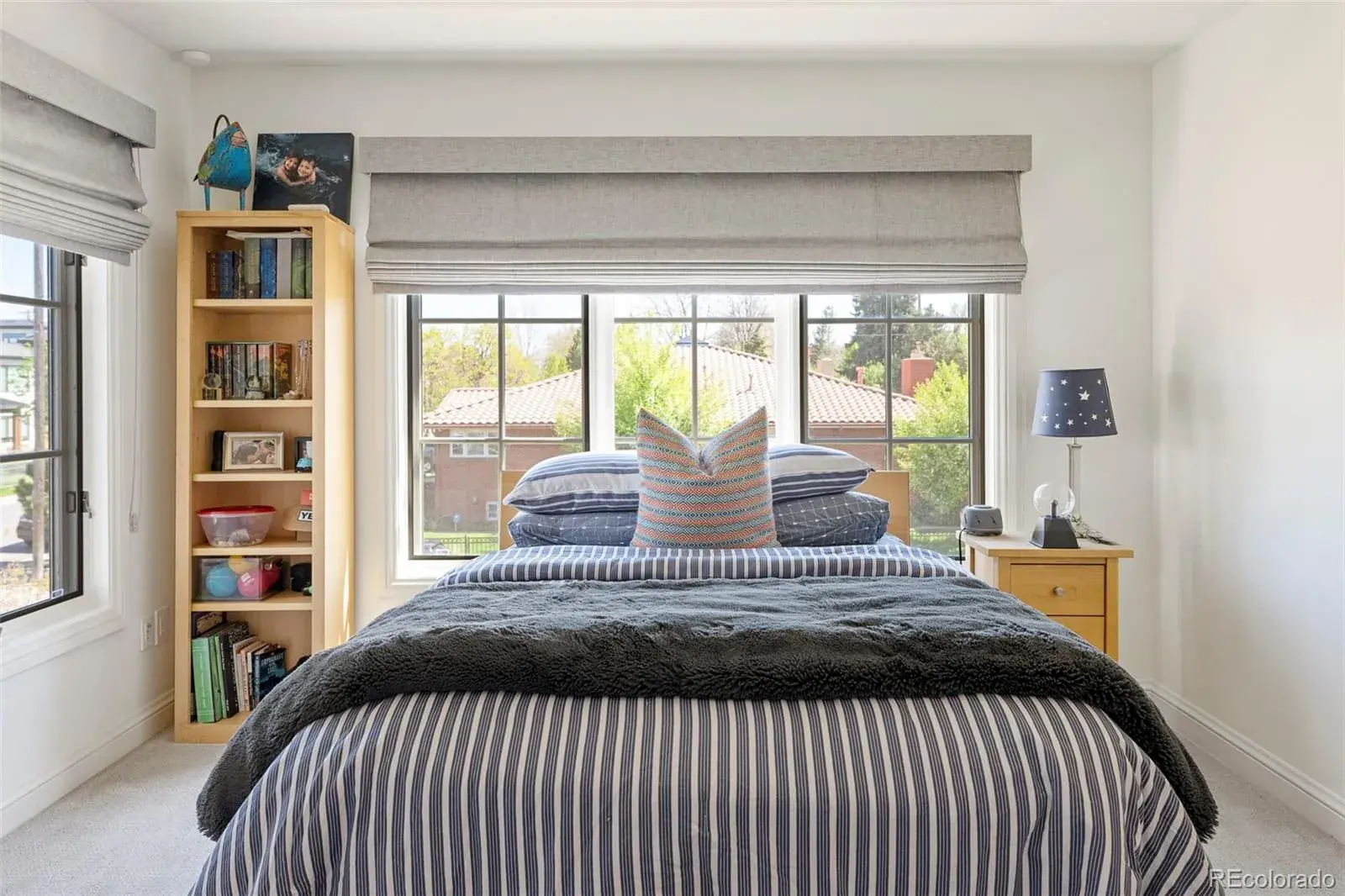 This is a cozy bedroom featuring a bed with striped bedding and a gray throw blanket. A window with gray roman shades provides natural light, and a bookshelf and nightstand with a lamp add to the room's functionality. The overall impression is comfortable and inviting.