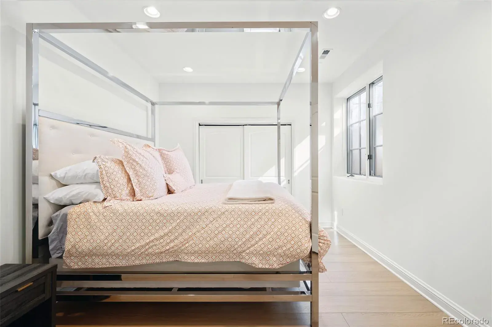 This is a bright and airy primary bedroom featuring a modern four-poster bed with a light-colored patterned duvet and several pillows. The room has white walls, light wood flooring, and a window providing natural light. The overall impression is clean, minimalist, and inviting.