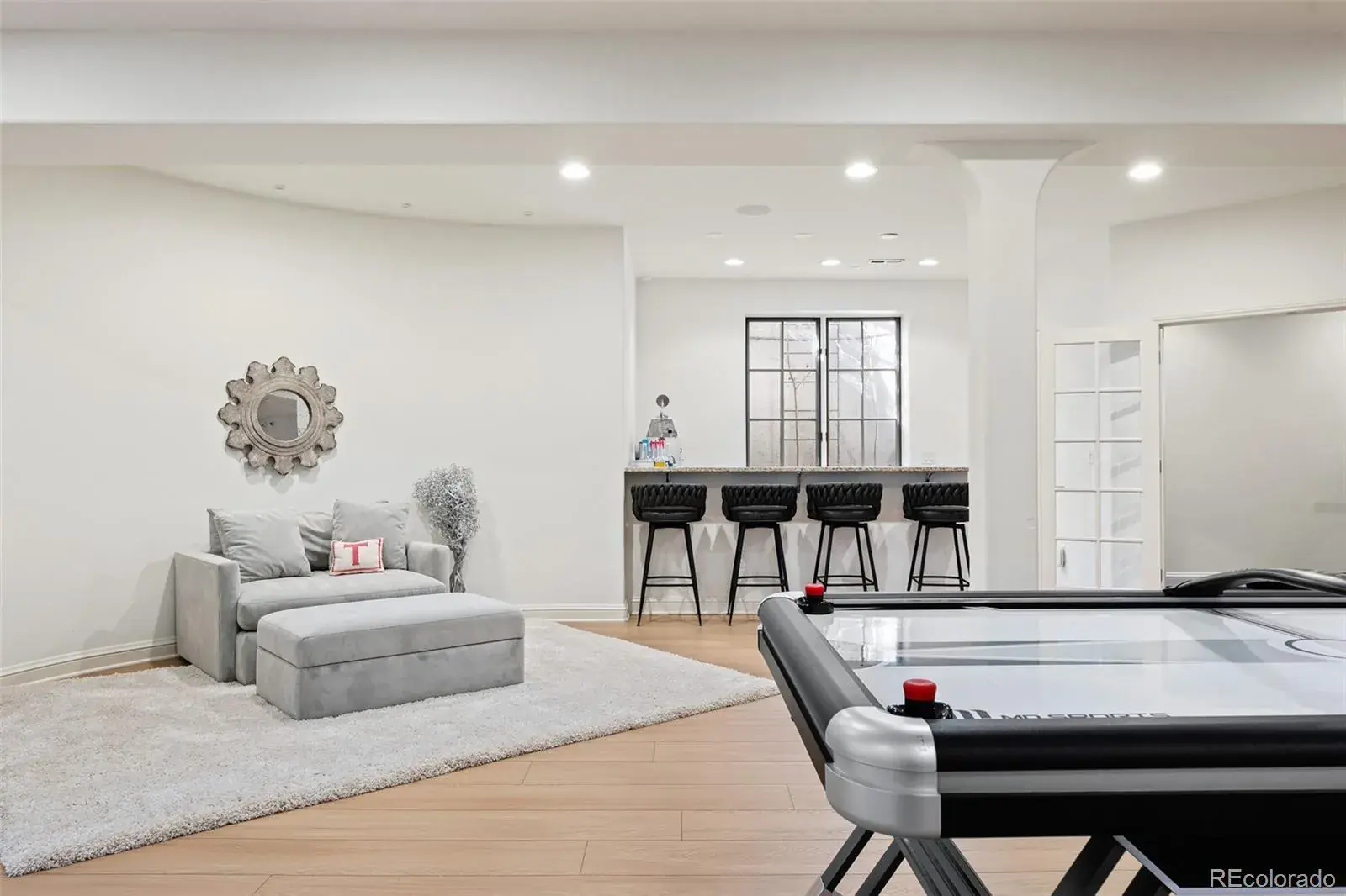 This is a well-lit basement area featuring a cozy seating area with a gray sofa and ottoman, complemented by a decorative mirror and a plush rug. In the background, there's a bar area with black stools and a window, and an air hockey table is prominently displayed in the foreground, suggesting a recreational space. The flooring is light wood, and the walls are painted in a neutral tone, creating a bright and inviting atmosphere.