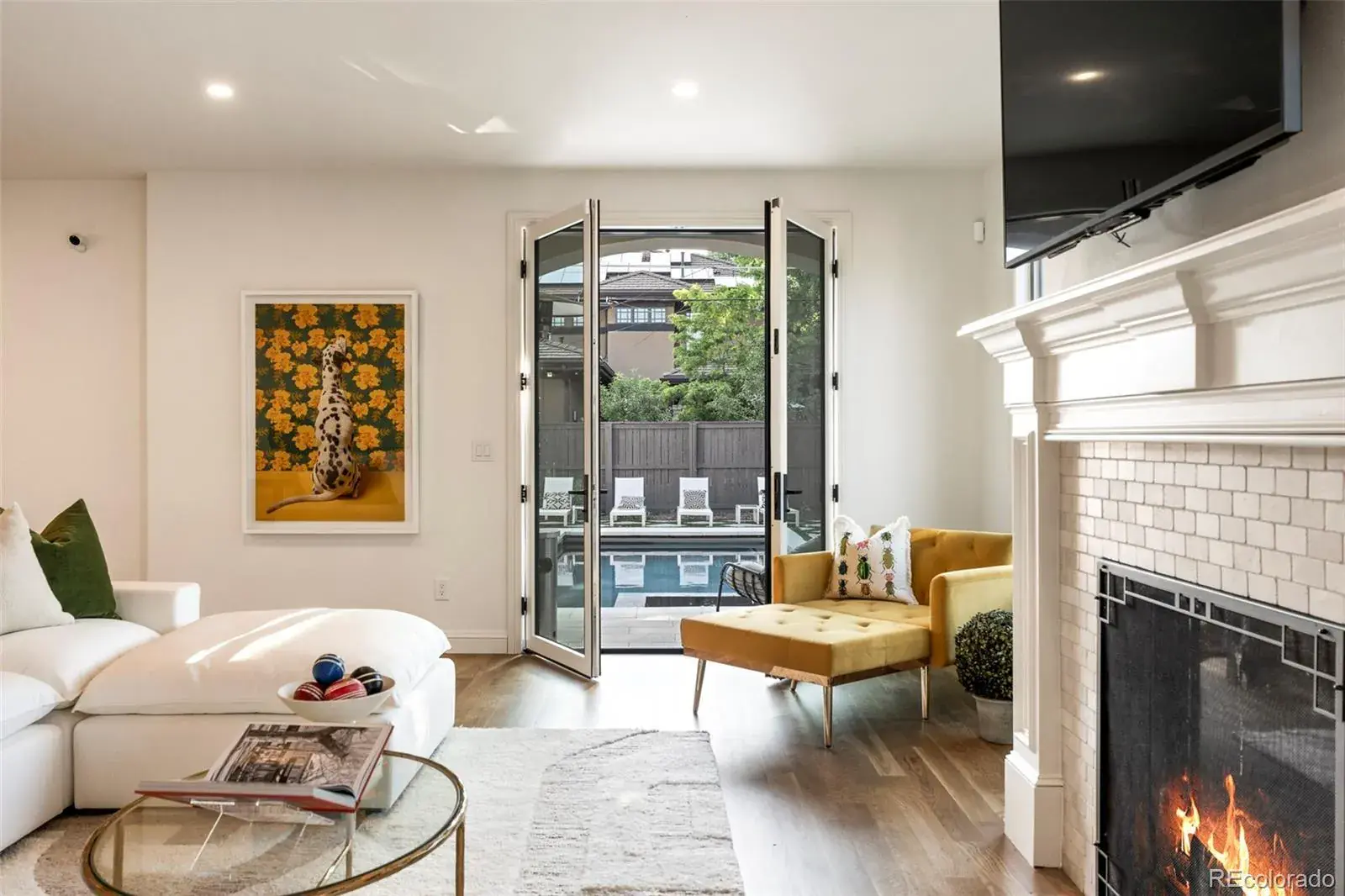 This is an interior shot of a living room featuring a modern design with hardwood floors and white walls. The room includes a white sectional sofa, a yellow chaise lounge, and a fireplace with a television mounted above it. French doors open to a view of a pool area, creating a bright and airy atmosphere.