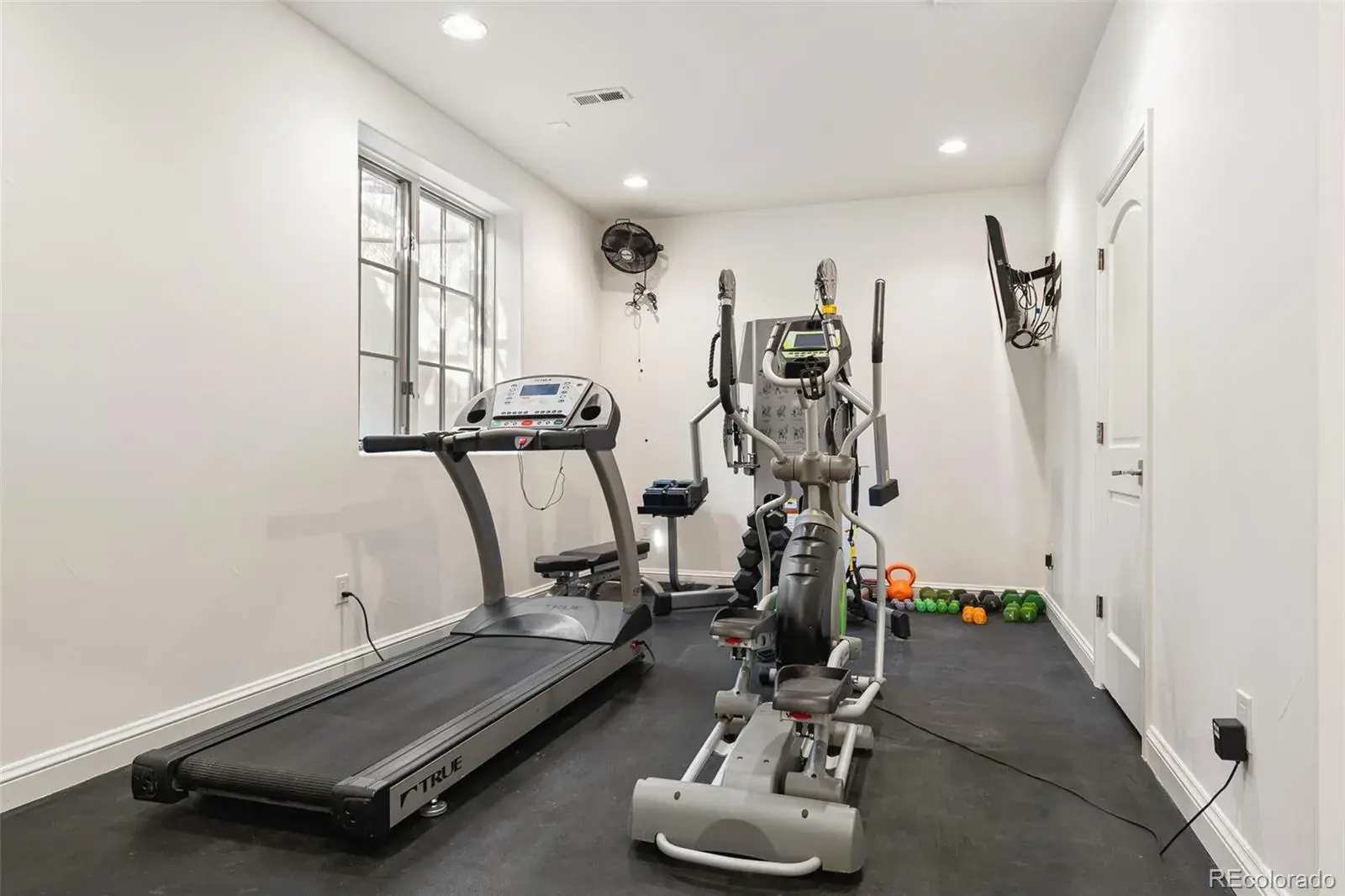 This is an interior shot of a home gym. The room features a treadmill, elliptical machine, and weight training equipment. The walls are painted white, and the floor is a dark rubber material. A window provides natural light, and a wall-mounted television is visible. The overall impression is a clean and functional workout space.