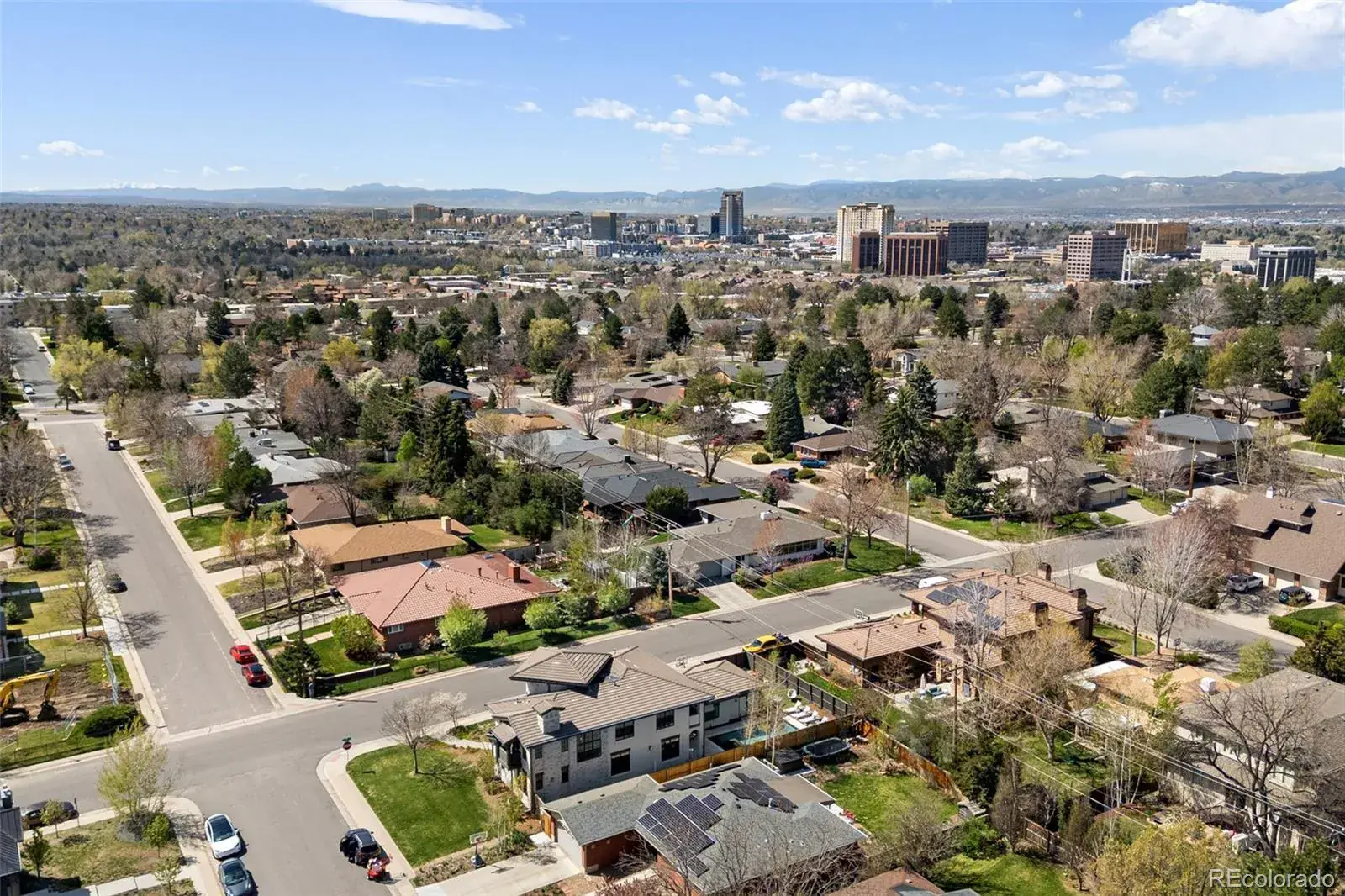 This aerial view showcases a residential neighborhood with a mix of single-family homes, mature trees, and well-maintained streets. The houses vary in style, some with red tile roofs and others with modern solar panels. In the background, a city skyline is visible, adding depth and context to the location.