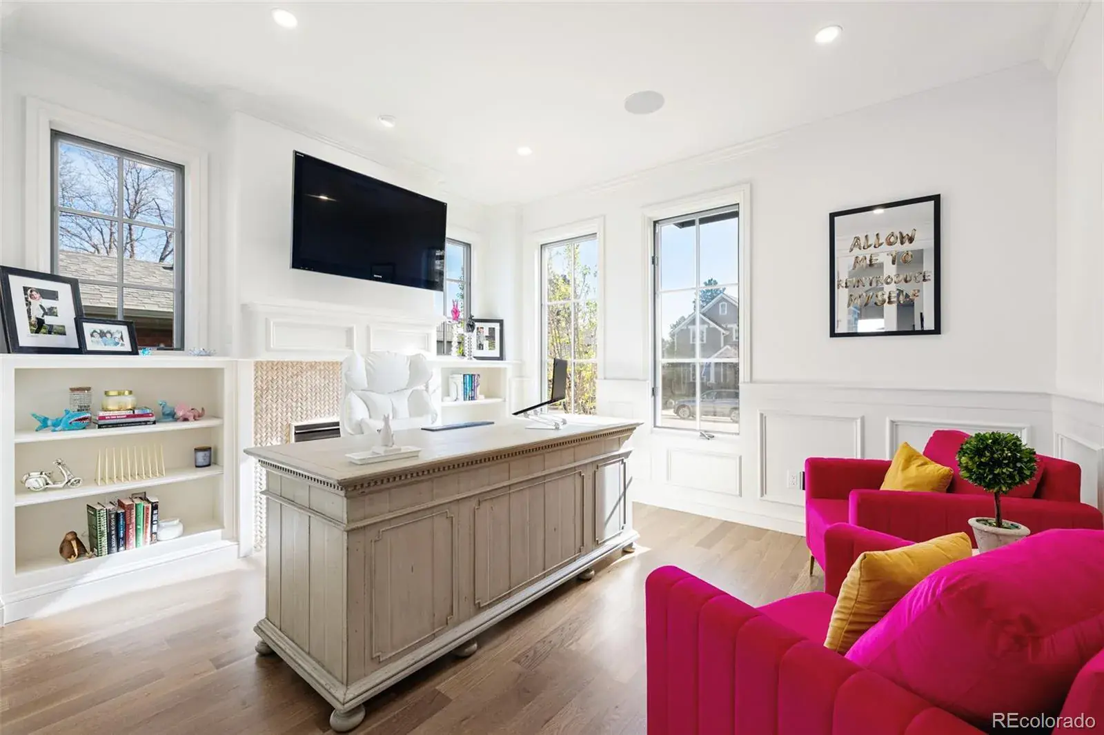 This is an interior shot of a well-lit home office featuring a large, distressed wood desk as the focal point. The room is painted in a bright white, complemented by white built-in bookshelves and wainscoting. Two vibrant pink armchairs with yellow pillows add a pop of color, creating a stylish and inviting workspace.