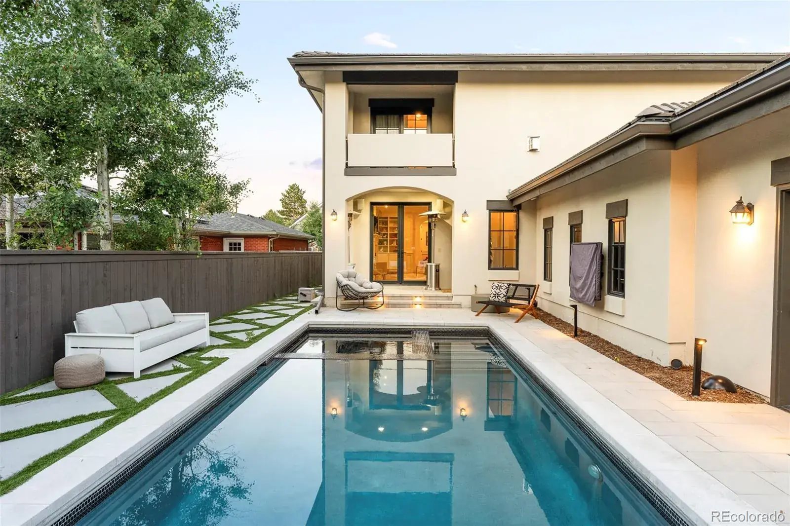 This image showcases a luxurious backyard featuring a rectangular pool with a dark tile edge, surrounded by light-colored paving stones and patches of grass. A modern white sofa sits alongside the pool, and the house's rear facade includes a balcony and arched doorway, creating an inviting outdoor living space. The pool's reflection adds depth and enhances the overall aesthetic.