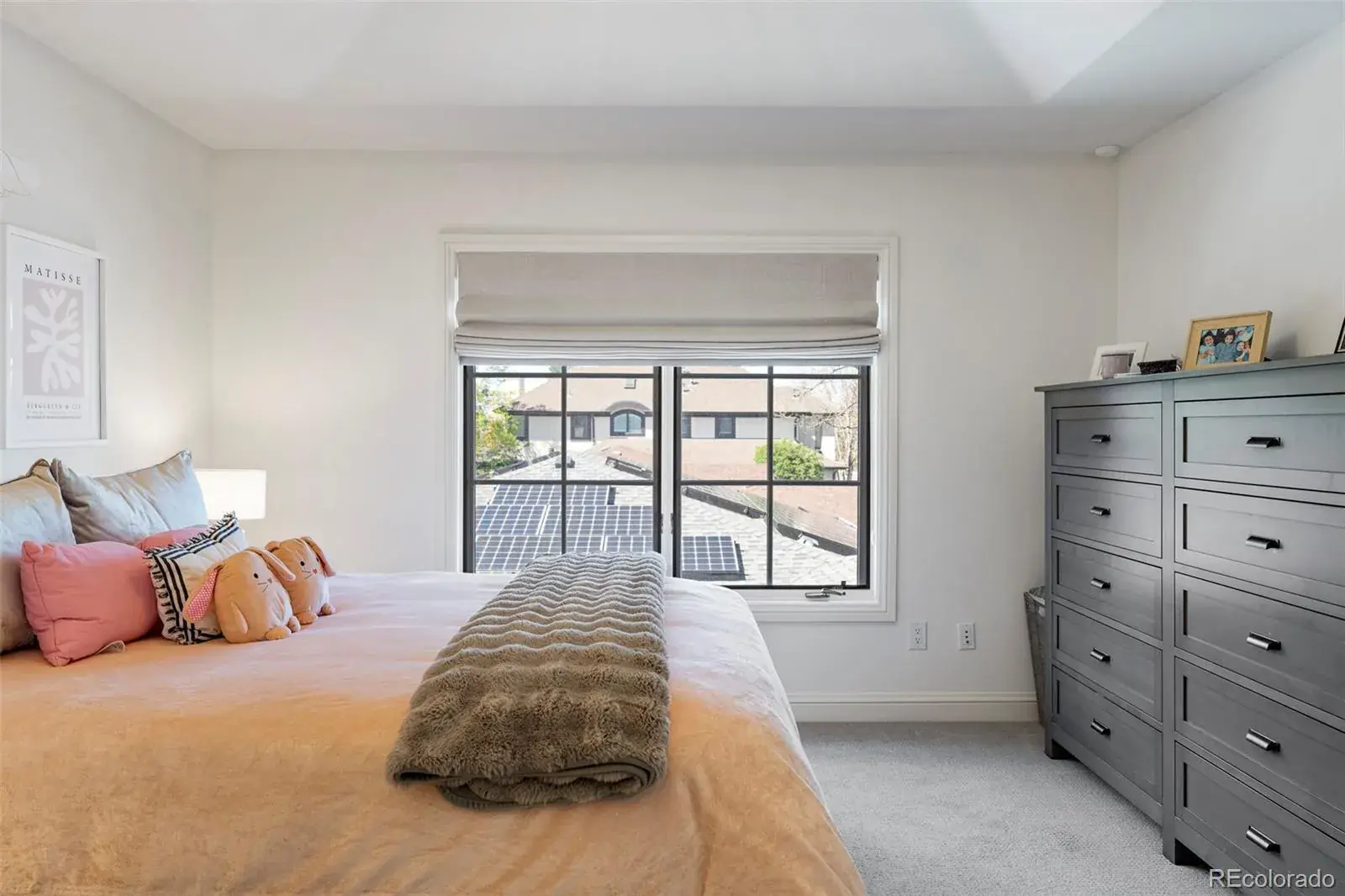This is a bright and inviting bedroom featuring a large window with a view of a neighboring house with solar panels. The room is decorated in neutral tones with a gray dresser and a bed adorned with plush pillows and a cozy throw. The overall impression is clean, comfortable, and well-lit, making it an appealing space.