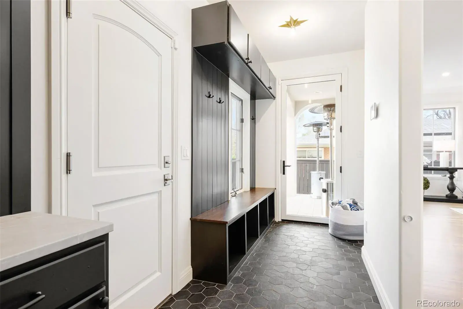 This interior shot showcases a stylish entryway with dark hexagonal tile flooring and a built-in mudroom bench with storage cubbies. A white door and a glass exterior door provide access, while a dark-toned cabinet and coat rack offer practical storage solutions. The space is well-lit and presents a clean, modern aesthetic.