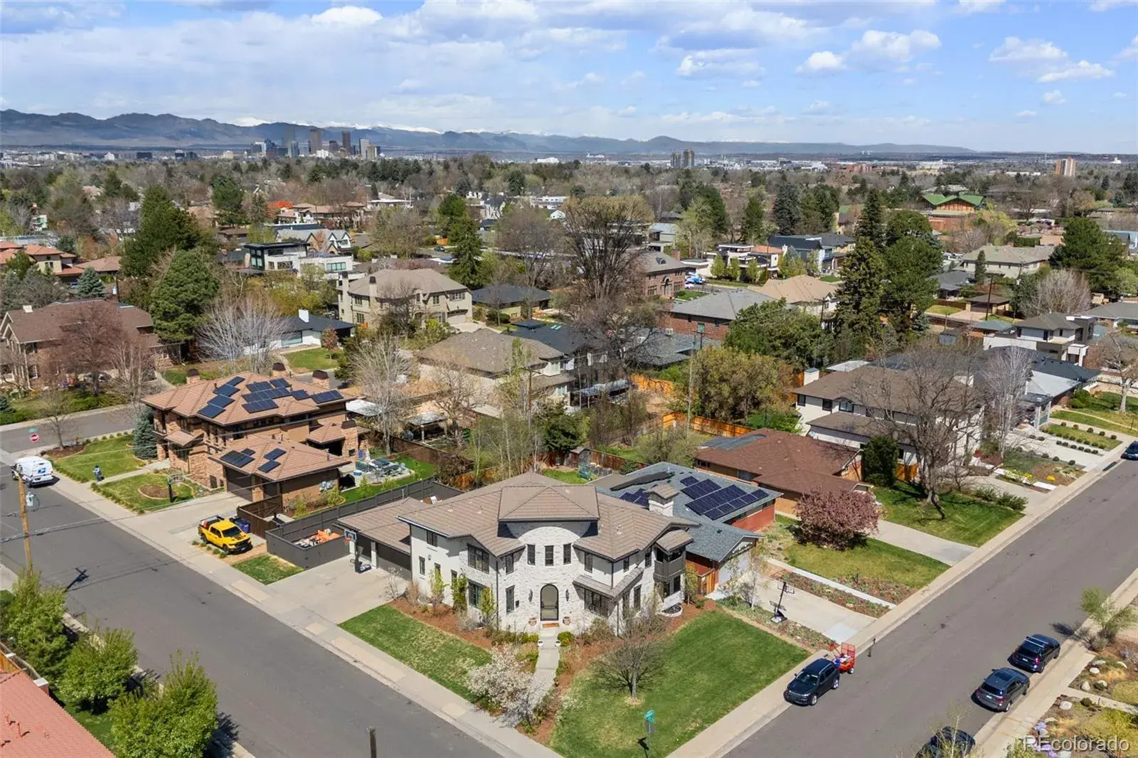This aerial shot showcases a neighborhood with well-maintained homes, mature trees, and manicured lawns. The focal point is a large, two-story house with a stone facade and a tile roof, surrounded by lush greenery. In the background, a cityscape and mountain range add depth and visual interest, suggesting a desirable location.
