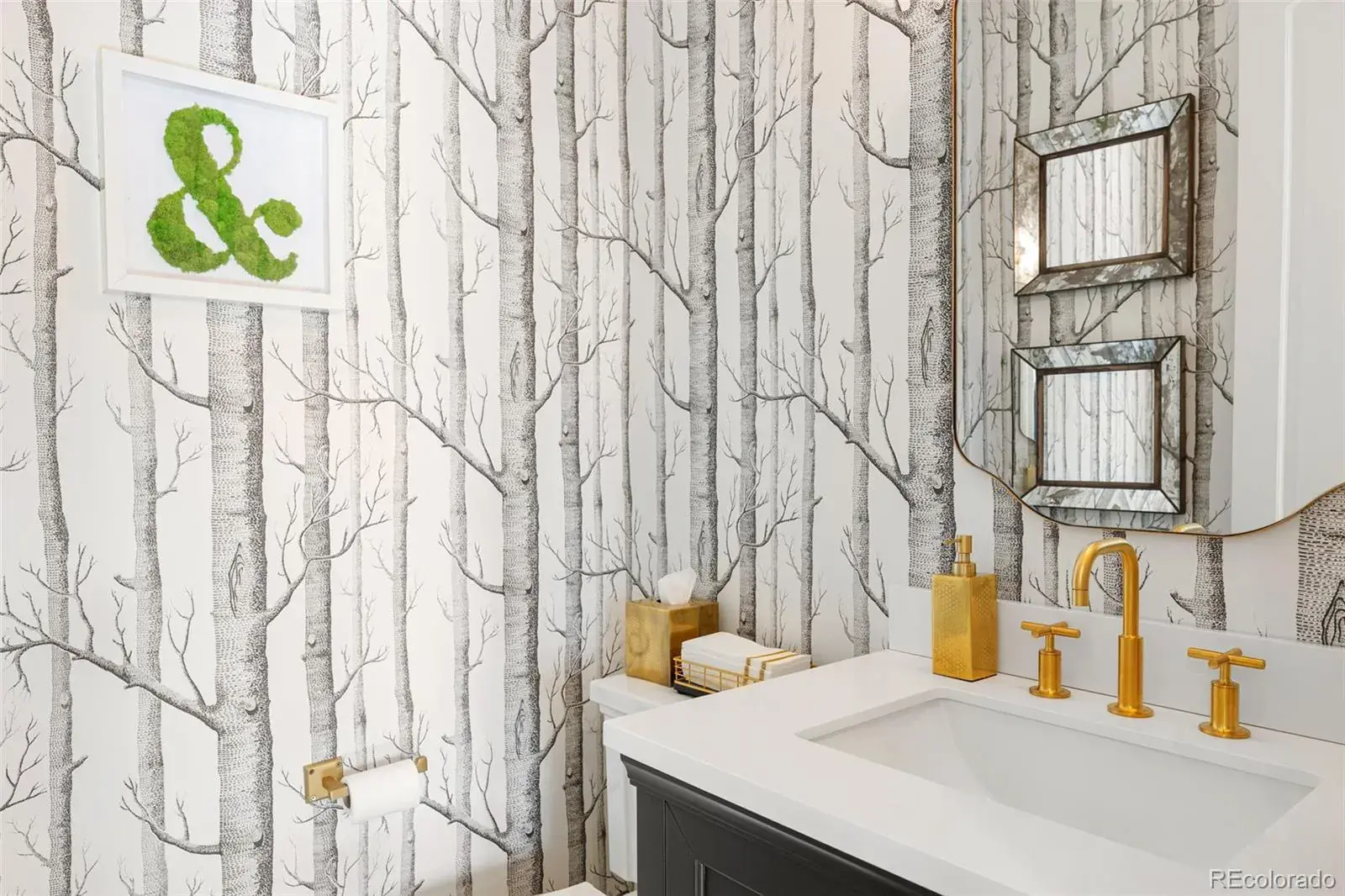 This is a stylish guest bathroom featuring a unique birch tree wallpaper that adds a touch of nature. The vanity has a dark base with a white countertop, complemented by gold fixtures and accessories. A framed moss ampersand adds a pop of color and whimsy to the space.