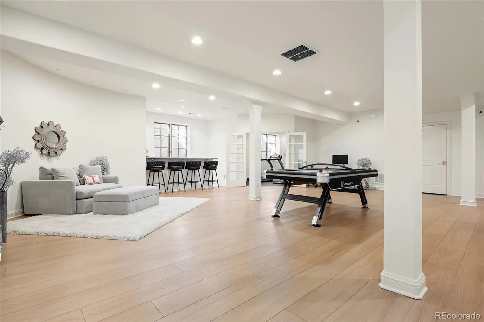 This is a spacious basement featuring light wood flooring and white walls, creating a bright and open atmosphere. The area includes a seating area with a gray sectional sofa and ottoman, a bar with black stools, and a pool table. Natural light filters through windows, enhancing the inviting feel of the space.