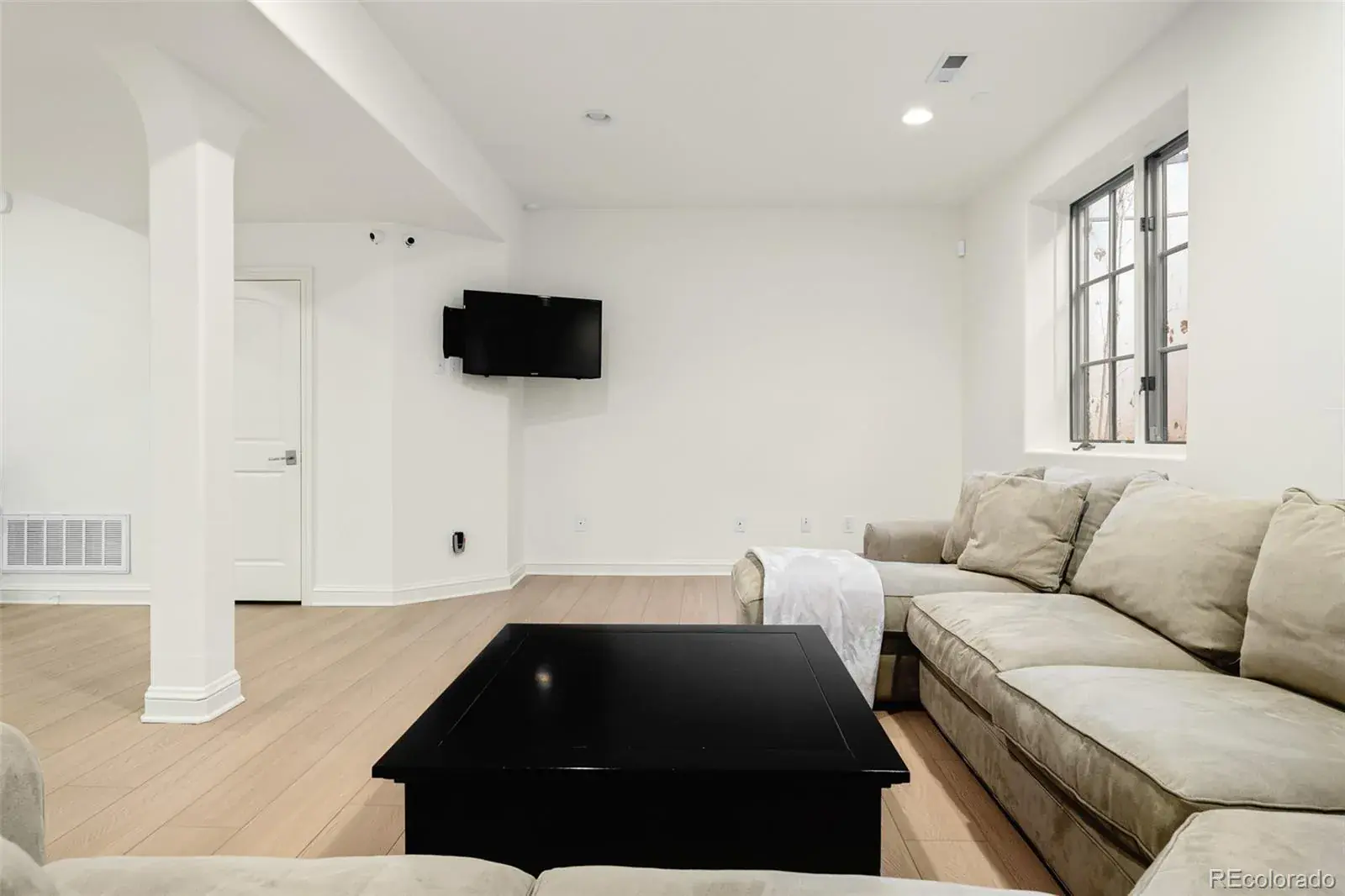 This is an interior shot of a living room featuring a large, comfortable sectional sofa in a neutral tone, a black coffee table, and a wall-mounted television. The room is well-lit with natural light coming through a window, and the walls are painted in a clean, bright white. The flooring is light-colored wood, adding warmth to the space.