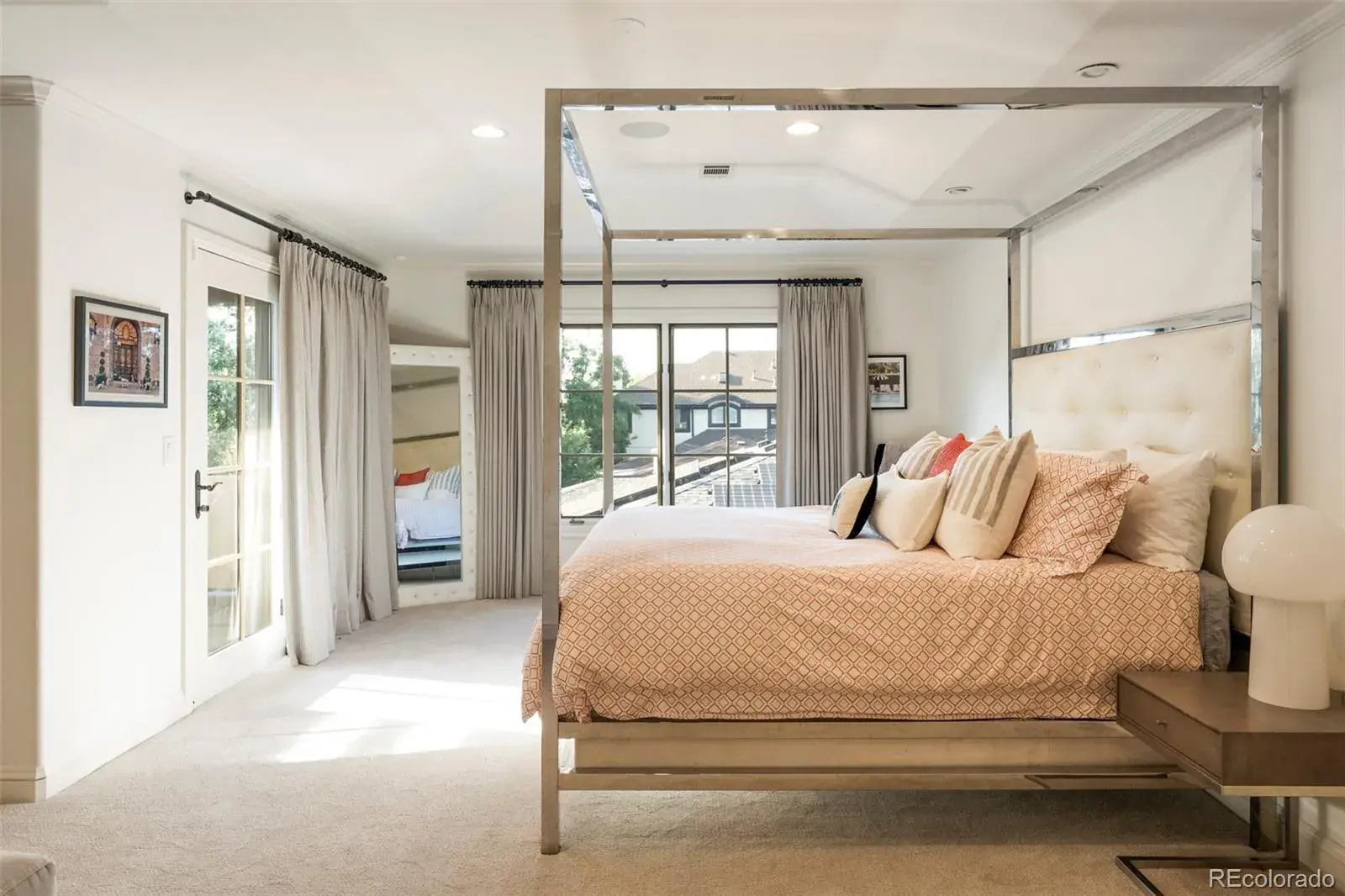 This is a bright and airy primary bedroom featuring a four-poster bed with a patterned duvet and multiple pillows. The room has neutral-toned walls, carpet, and curtains, with natural light streaming in through large windows. A mirrored closet door and a framed picture add to the room's decor.