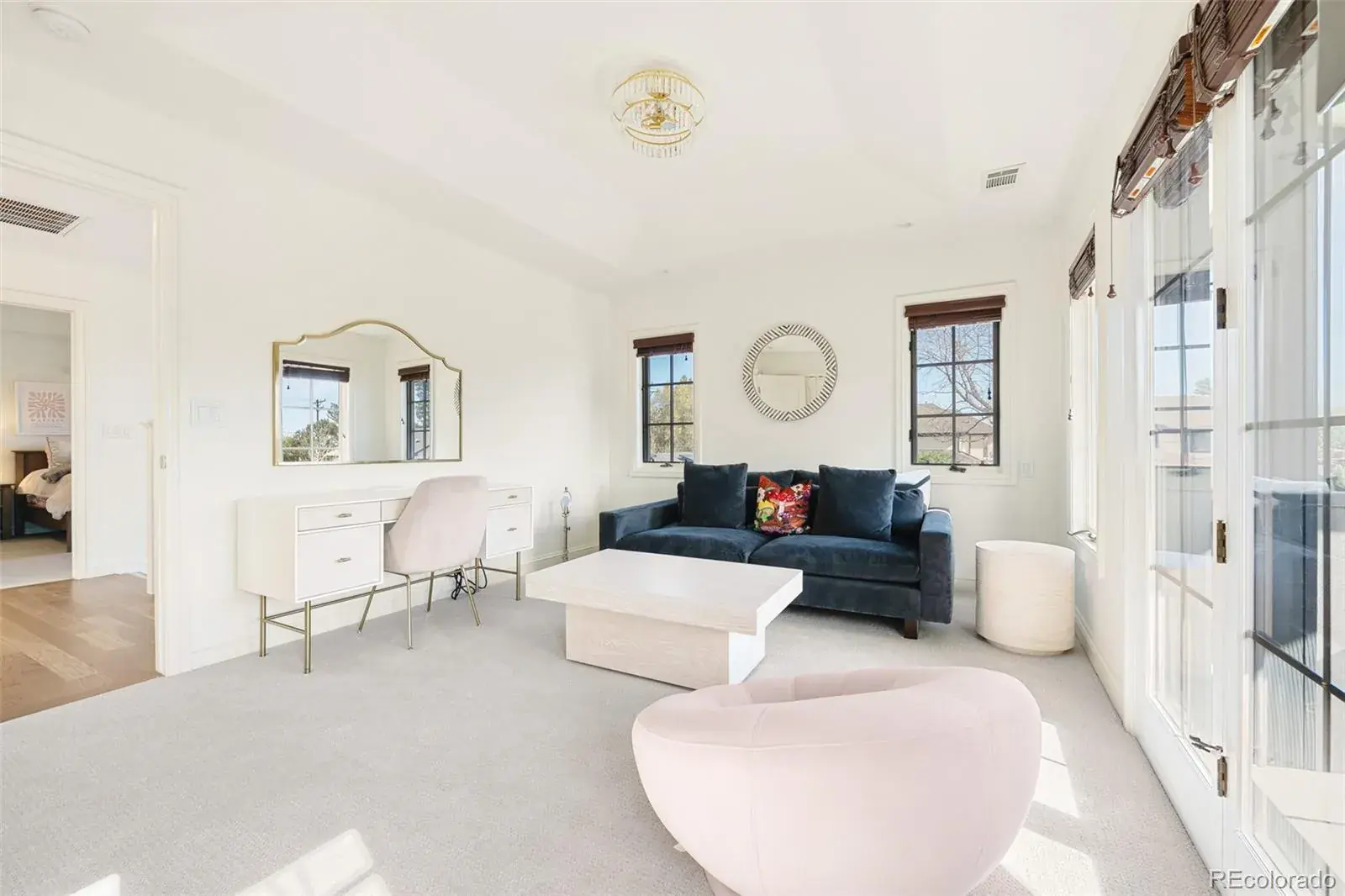 This is a bright and airy bedroom featuring white walls and light gray carpeting. The room is furnished with a blue velvet sofa, a white coffee table, and a vanity with a gold-framed mirror. Natural light floods the space through multiple windows and French doors, creating a welcoming and serene atmosphere.