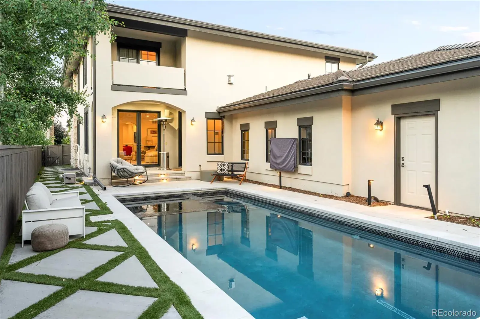 This image showcases a modern backyard featuring a rectangular pool with a sleek, tiled edge. The pool is surrounded by a concrete patio interspersed with geometric patches of grass, creating a contemporary design. The exterior of the house is visible, featuring a light-colored stucco finish with dark trim, adding to the sophisticated and inviting atmosphere.