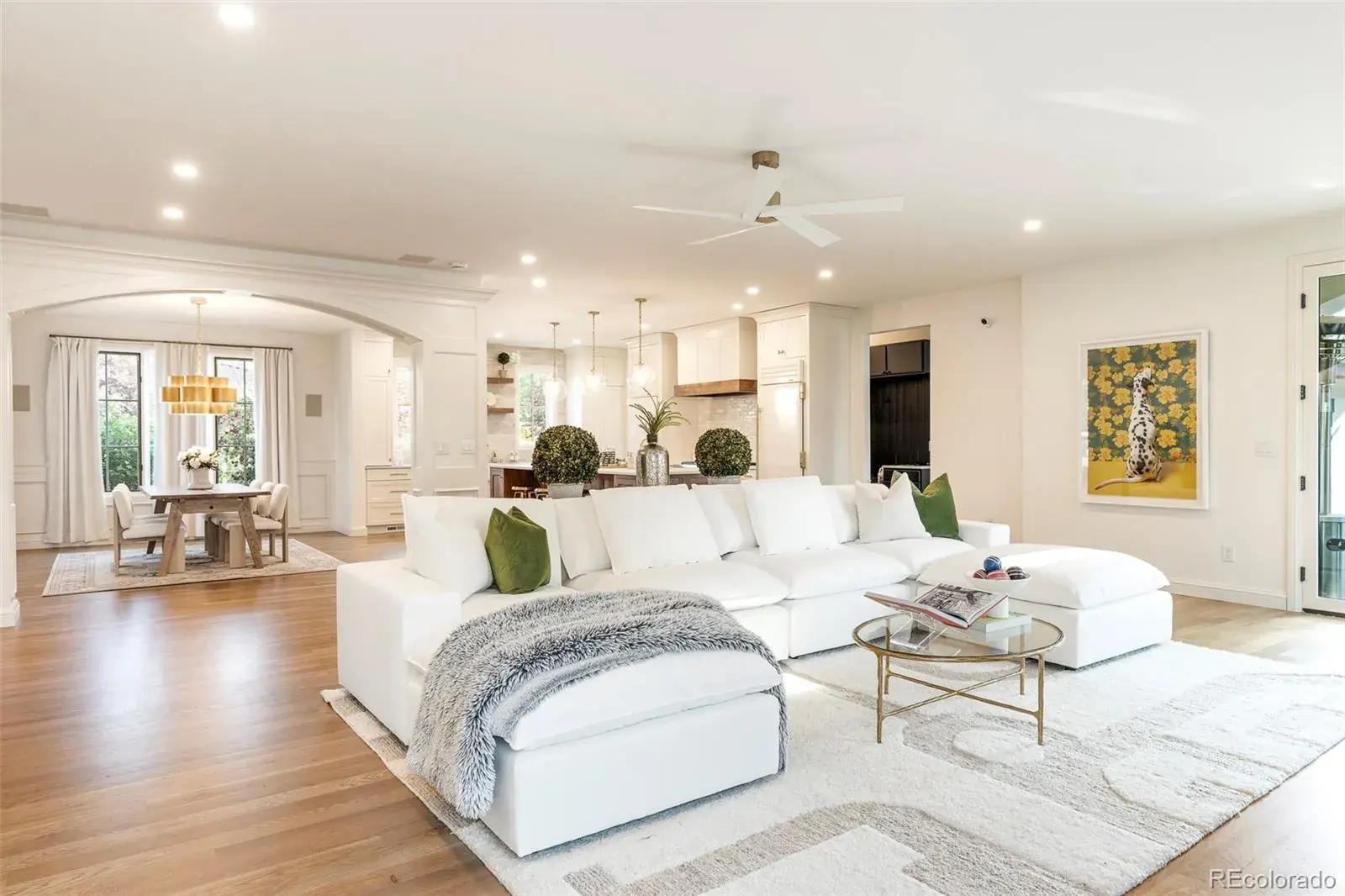 This is a bright and airy living room featuring an open-concept design that seamlessly connects to the dining area and kitchen. The focal point is a large, white sectional sofa complemented by a neutral-toned rug and a glass-topped coffee table. The space is well-lit with recessed lighting and natural light, creating a welcoming and modern atmosphere.
