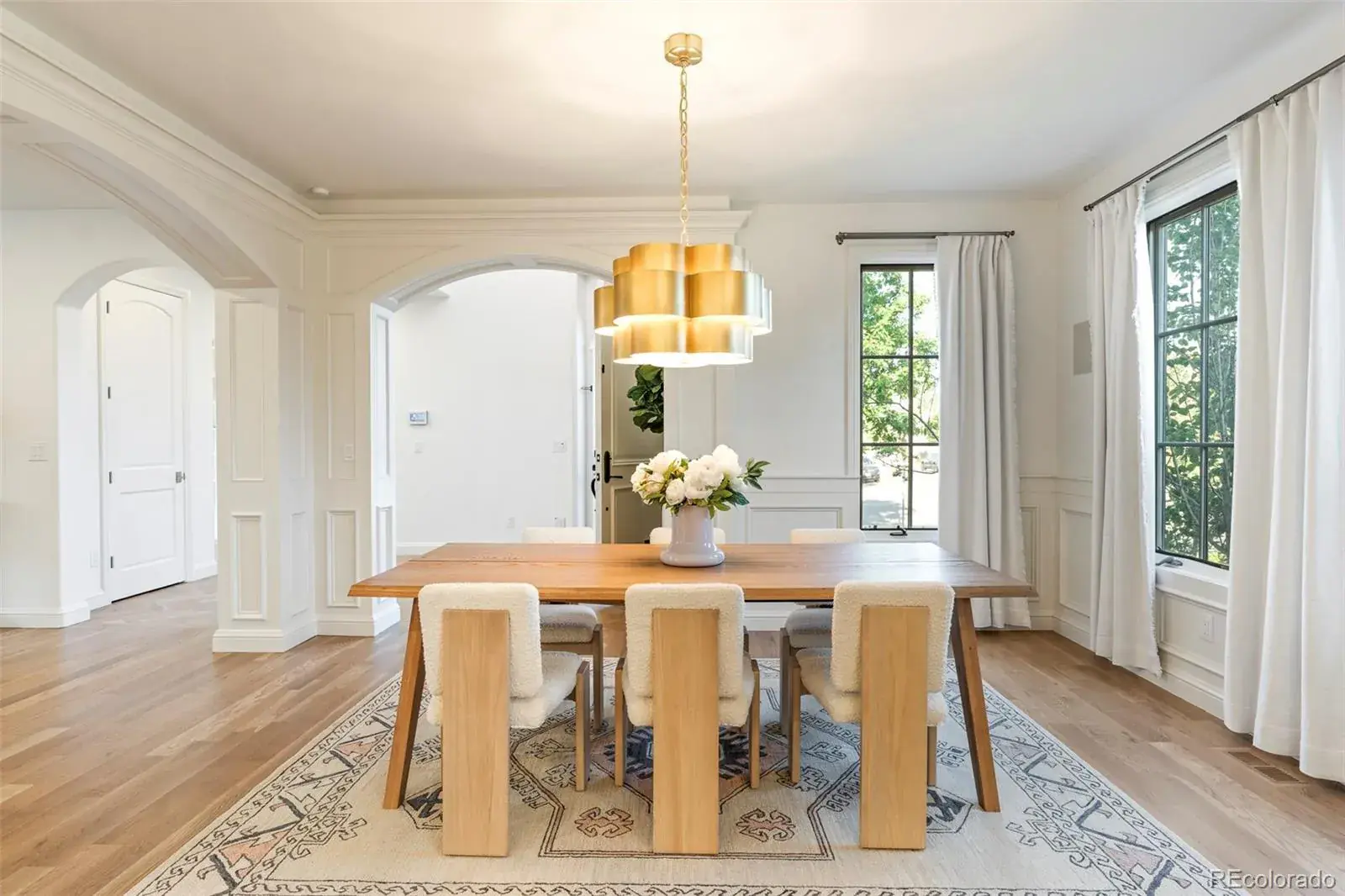 This is an interior shot of a dining room featuring a wooden table with six chairs, a modern gold chandelier, and a patterned rug. The room has white walls, trim, and curtains, with natural light coming in from the windows. The overall style is elegant and inviting.