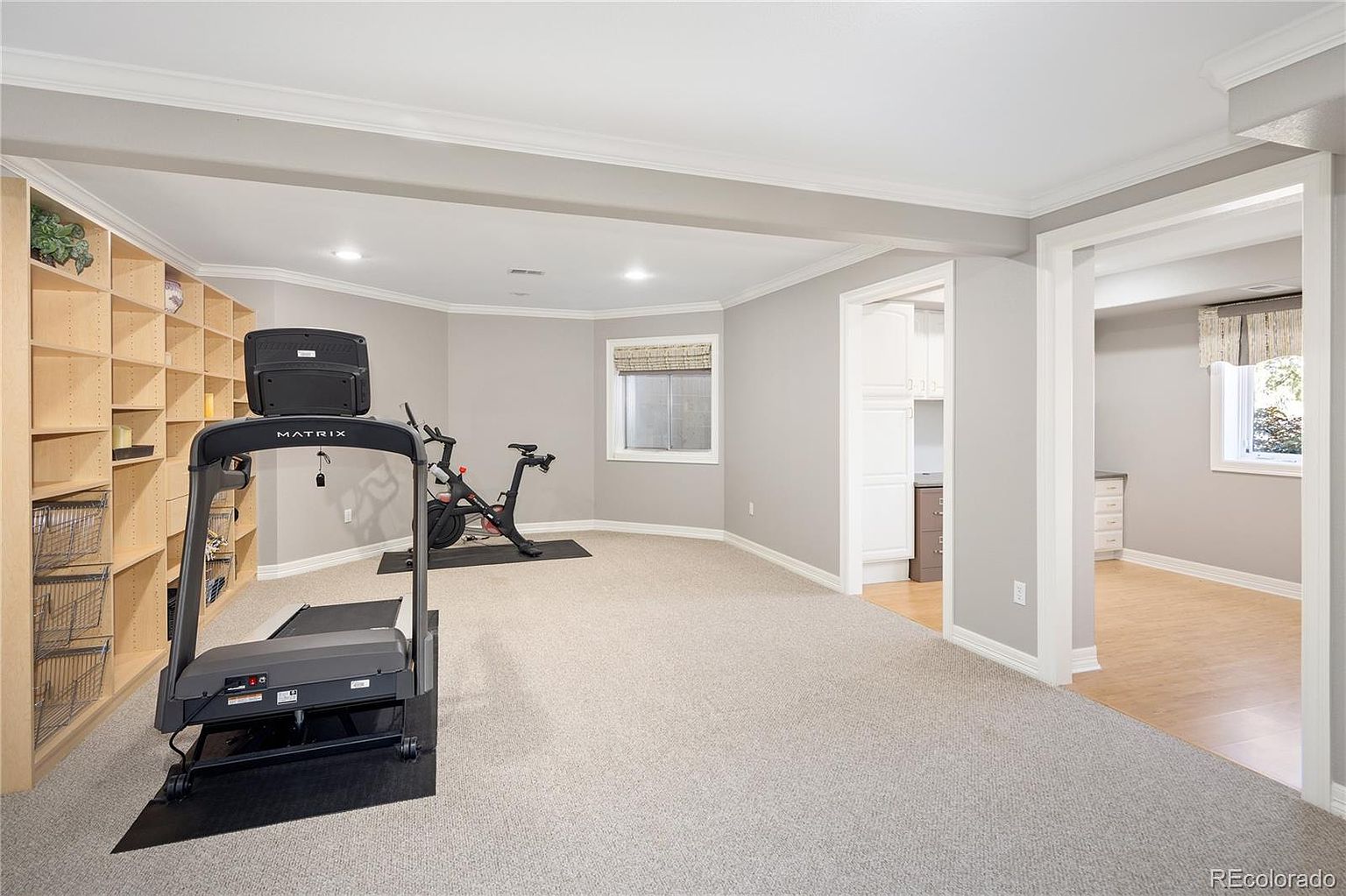This home gym features a neutral color palette with light gray walls and plush carpeting, creating a clean and functional workout space. The room is equipped with a Matrix treadmill and a stationary bike, complemented by extensive built-in shelving for storage. A wide doorway leads into an adjacent room with light wood flooring, providing an open and airy feel to the lower-level living area.