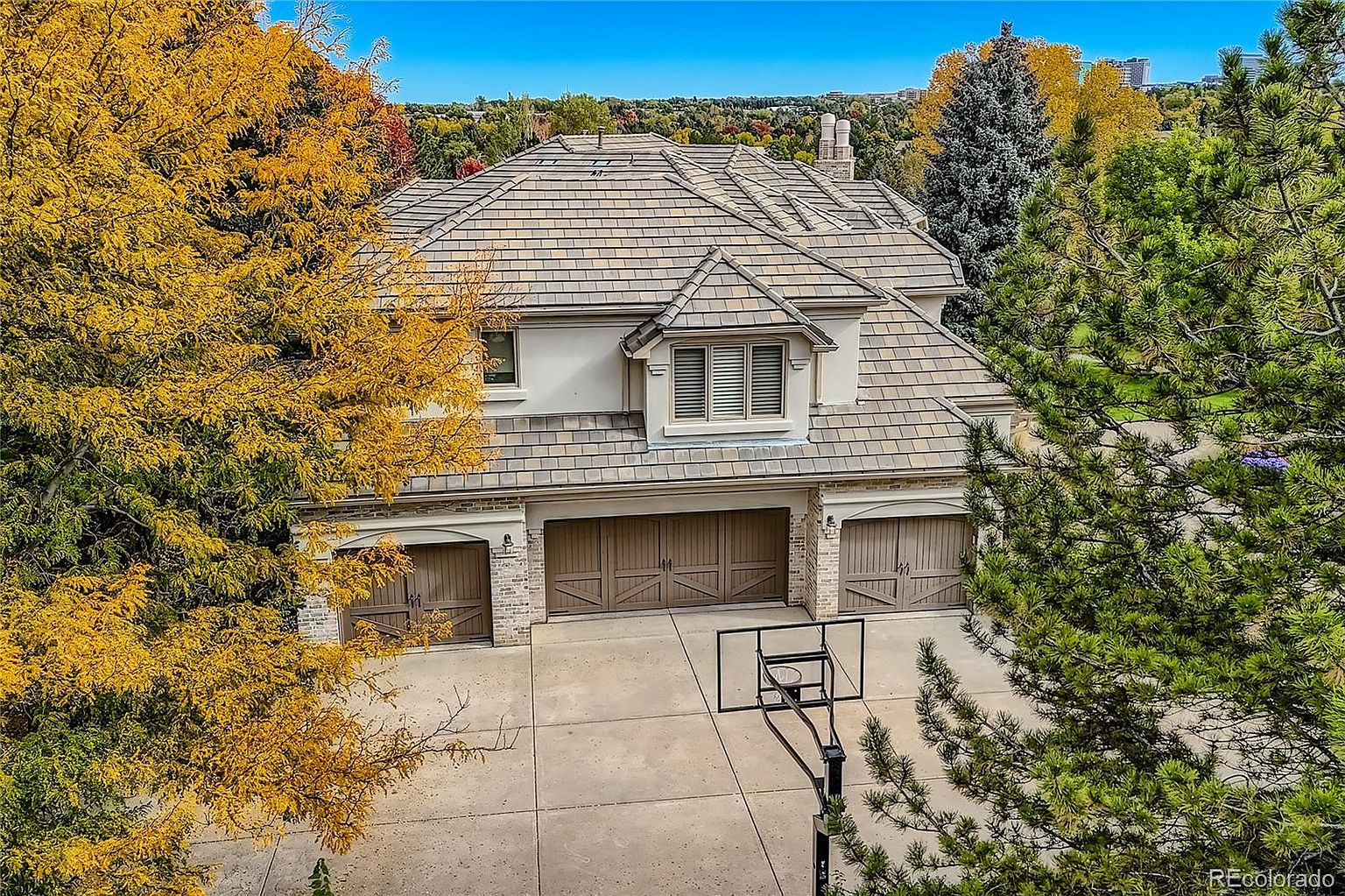 This high-angle aerial shot captures the front facade of a stately, multi-story home featuring a three-car garage with carriage-style doors. The exterior is finished in a light-colored stucco with brick accents, complemented by a complex, multi-gabled roofline. A basketball hoop stands on the spacious concrete driveway, framed by vibrant autumn foliage and mature evergreen trees, creating a picturesque and inviting suburban setting.