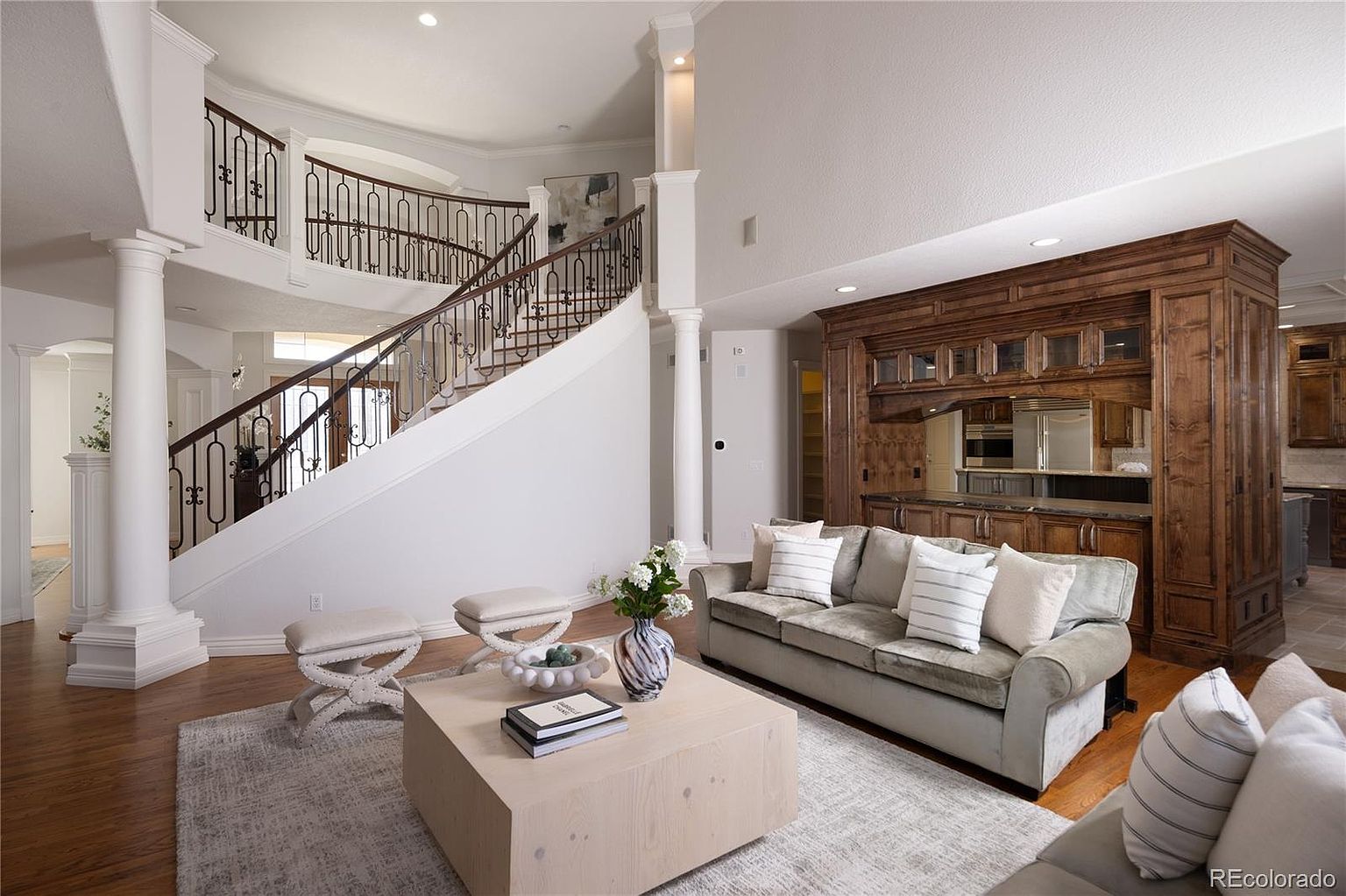 This elegant, high-ceilinged living room features a grand curved staircase with ornate iron railings as a central architectural focal point. The space is anchored by a plush, light-colored sofa and a large, minimalist wooden coffee table, complemented by a built-in dark wood cabinetry unit that adds warmth and sophistication. The open-concept design flows seamlessly into the background, creating an airy and luxurious atmosphere perfect for entertaining.