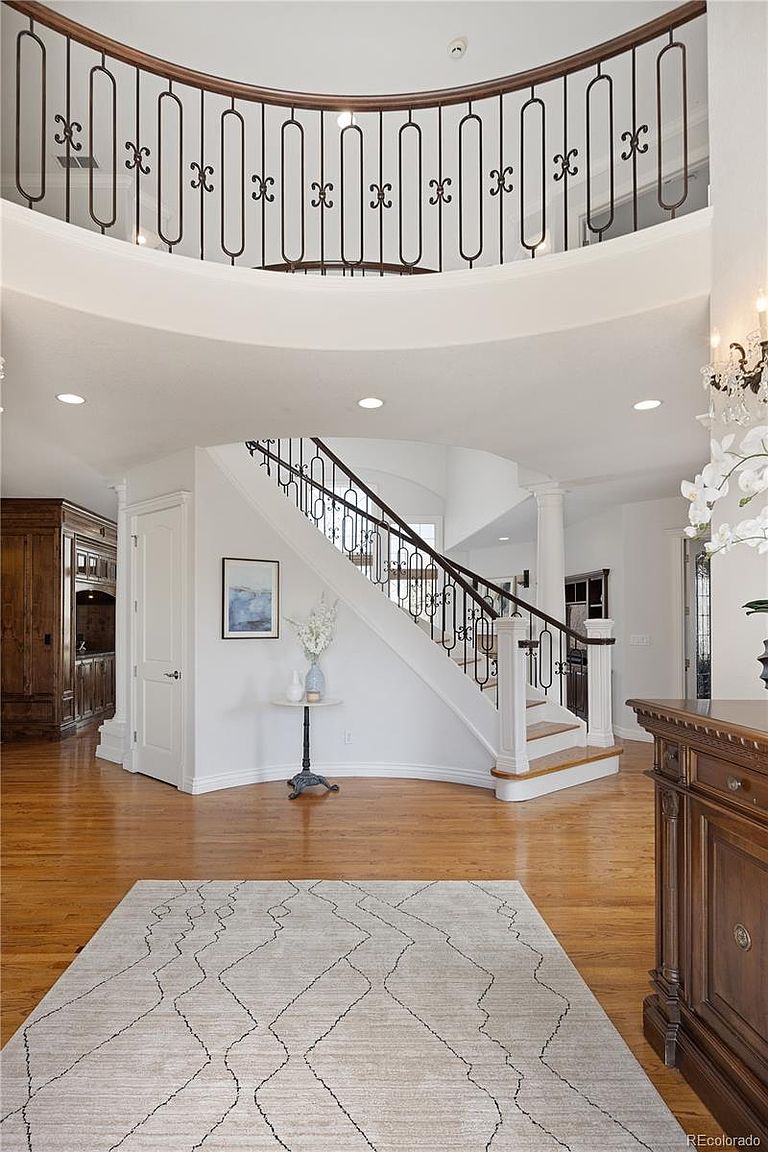 This grand foyer features a sweeping curved staircase with intricate wrought-iron railings and a matching balcony overlooking the entryway. The space is defined by warm hardwood flooring, a neutral color palette, and a sophisticated, open-concept design that creates an inviting and luxurious atmosphere. A minimalist area rug centers the room, while elegant architectural details and recessed lighting enhance the home's high-end aesthetic.