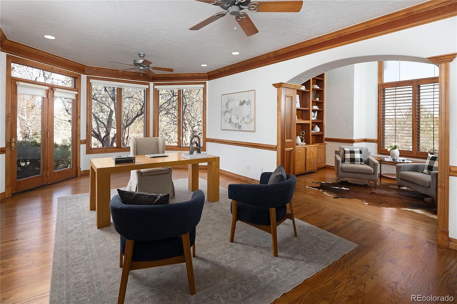 This spacious and elegant home office features rich hardwood flooring, custom wood trim, and a large desk positioned to take advantage of natural light from the surrounding windows. The room is divided by a graceful archway, leading to a cozy seating area with two armchairs and a bookshelf, creating a sophisticated and functional workspace. The overall aesthetic is traditional and warm, offering a professional yet inviting atmosphere.