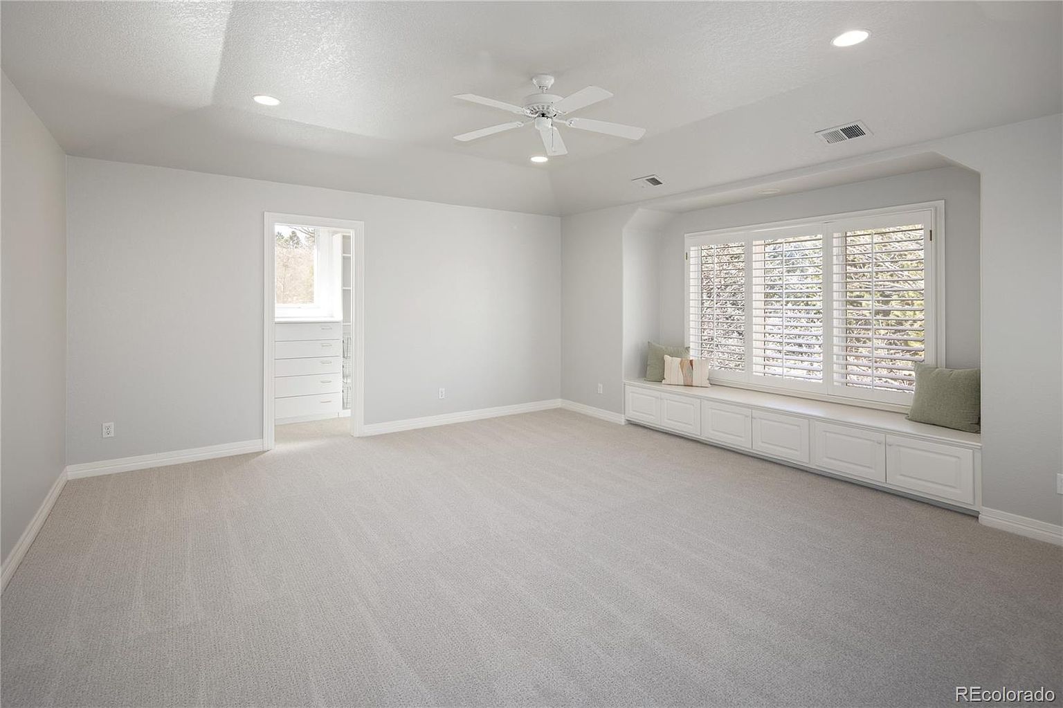 This spacious primary bedroom features neutral gray walls, plush carpeting, and a vaulted ceiling with a central white ceiling fan. A large window seat with built-in storage cabinets provides a cozy nook beneath expansive windows, while an open doorway leads into a walk-in closet. The room offers a clean, bright, and airy atmosphere, perfect for a tranquil retreat.