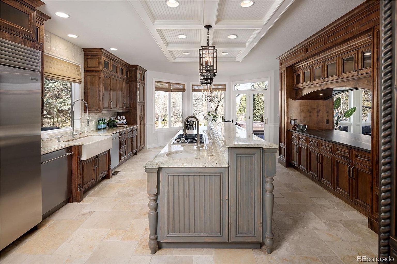 This expansive, high-end kitchen features rich, dark-stained wood cabinetry contrasted by a central island with a distressed grey finish and a light granite countertop. The space is illuminated by recessed lighting and a decorative chandelier, while a large farmhouse sink and stainless steel appliances add both charm and functionality. The perspective is a wide, centered shot that highlights the symmetrical layout and the seamless flow between the cooking area and the bright, window-filled breakfast nook.