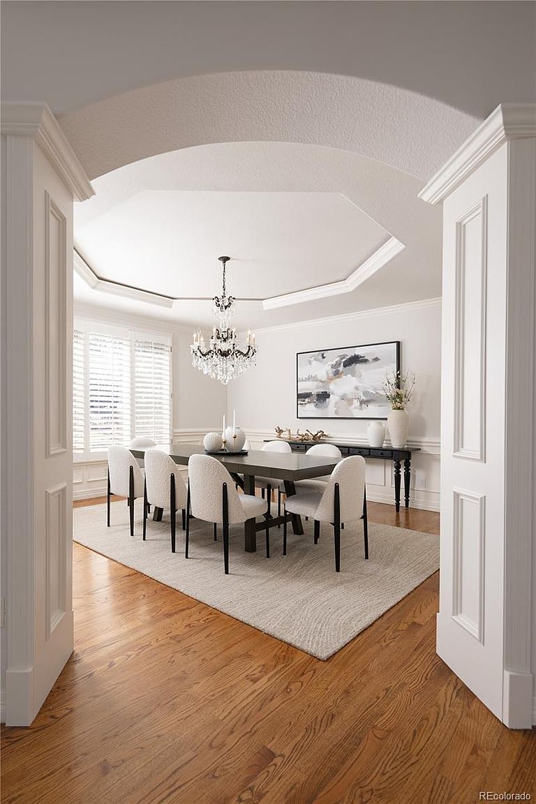 This elegant dining room is framed by a grand arched entryway, showcasing a sophisticated space with a dark wood table surrounded by modern, cream-colored upholstered chairs. The room features a striking octagonal tray ceiling with a crystal chandelier, hardwood flooring, and a minimalist console table adorned with decor. The overall aesthetic is refined and bright, offering a perfect setting for formal gatherings.