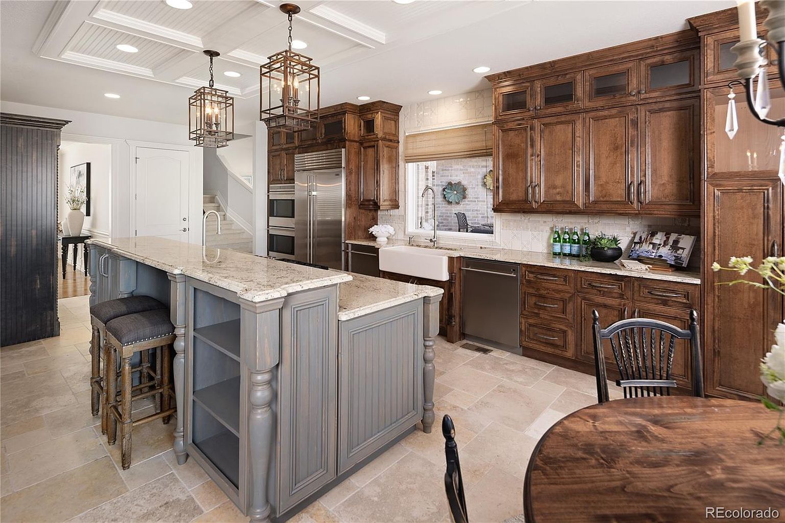 This elegant, traditional-style kitchen features rich, dark-stained wood cabinetry contrasted by a large, grey-painted center island with a granite countertop. The space is well-lit by decorative lantern-style pendant lights and recessed lighting, highlighting the stone tile flooring and farmhouse sink. A dining area is visible in the foreground, creating an inviting and functional layout perfect for family gatherings.