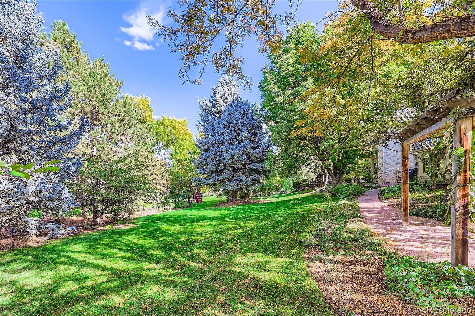 This expansive, lush backyard features a well-manicured lawn surrounded by mature trees, including striking blue spruces and deciduous trees with hints of autumn color. A brick pathway leads along the side of the home, partially shaded by a rustic wooden pergola structure. The scene offers a serene and private atmosphere, perfect for outdoor relaxation or recreation.