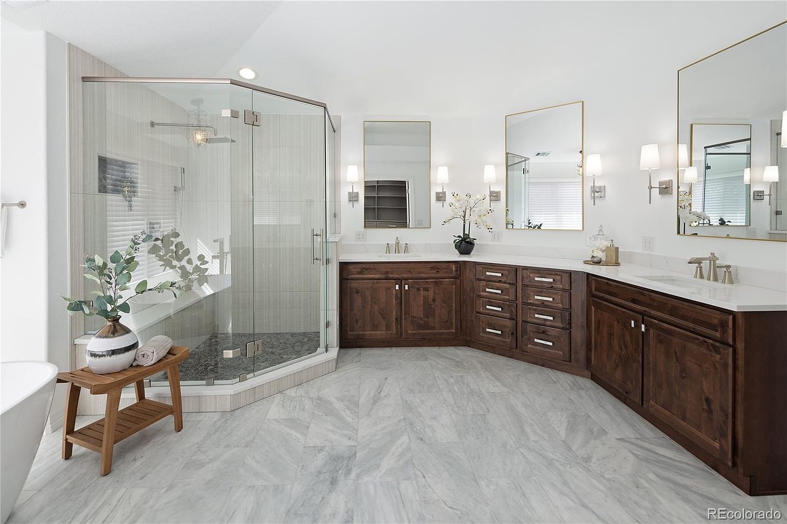 This spacious primary bathroom features a luxurious walk-in glass shower with stone tile flooring and a double vanity with rich, dark wood cabinetry topped with white countertops. The room is bright and airy, accented by gold-framed mirrors, elegant wall sconces, and large-format marble-look floor tiles. A wooden bench sits in the foreground, adding a warm, organic touch to the sophisticated, modern design.
