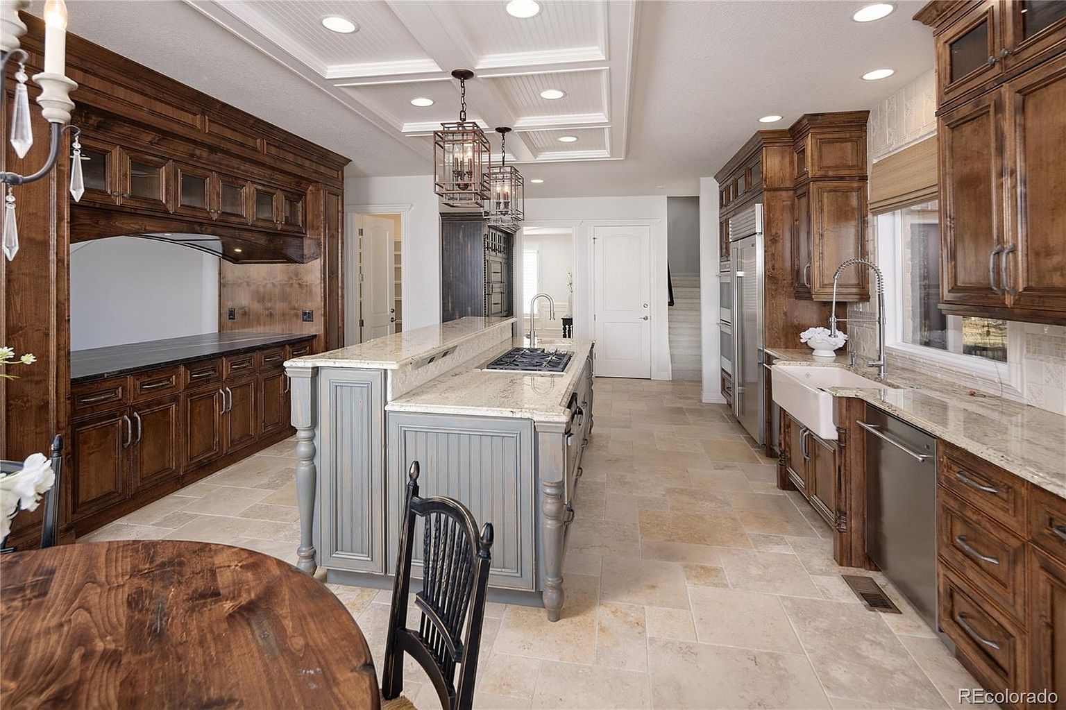 This spacious, high-end kitchen features rich, dark wood cabinetry contrasted by a large, light-grey central island with a granite countertop and integrated cooktop. The room is illuminated by elegant pendant lighting and recessed ceiling lights, highlighting the travertine tile flooring and coffered ceiling details. A farmhouse-style sink, stainless steel appliances, and a warm, traditional aesthetic create a sophisticated and inviting culinary space.