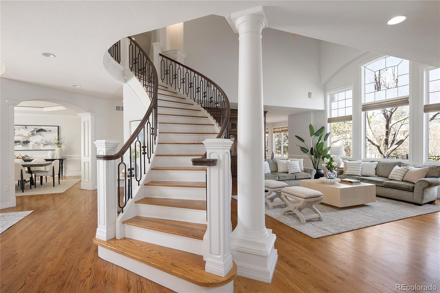 This elegant foyer features a grand, curved staircase with dark wood handrails and white balusters as the central focal point. To the right, an open-concept living area is bathed in natural light from large windows, while a formal dining room is visible to the left. The space is defined by high ceilings, hardwood flooring, and a classic architectural column, creating a sophisticated and welcoming atmosphere.