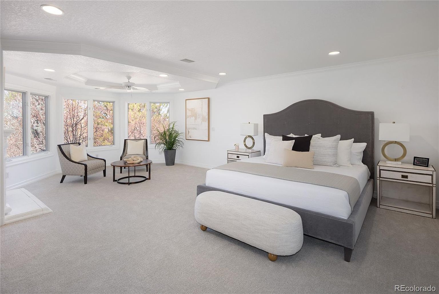 This spacious primary bedroom features a large, plush grey upholstered bed as the centerpiece, complemented by a matching bench and elegant nightstands. The room is bathed in natural light from a curved wall of windows that frame a serene outdoor view, creating a bright and airy atmosphere. A cozy seating area with two patterned armchairs and a small coffee table adds a touch of luxury and comfort to this expansive, neutrally-toned suite.
