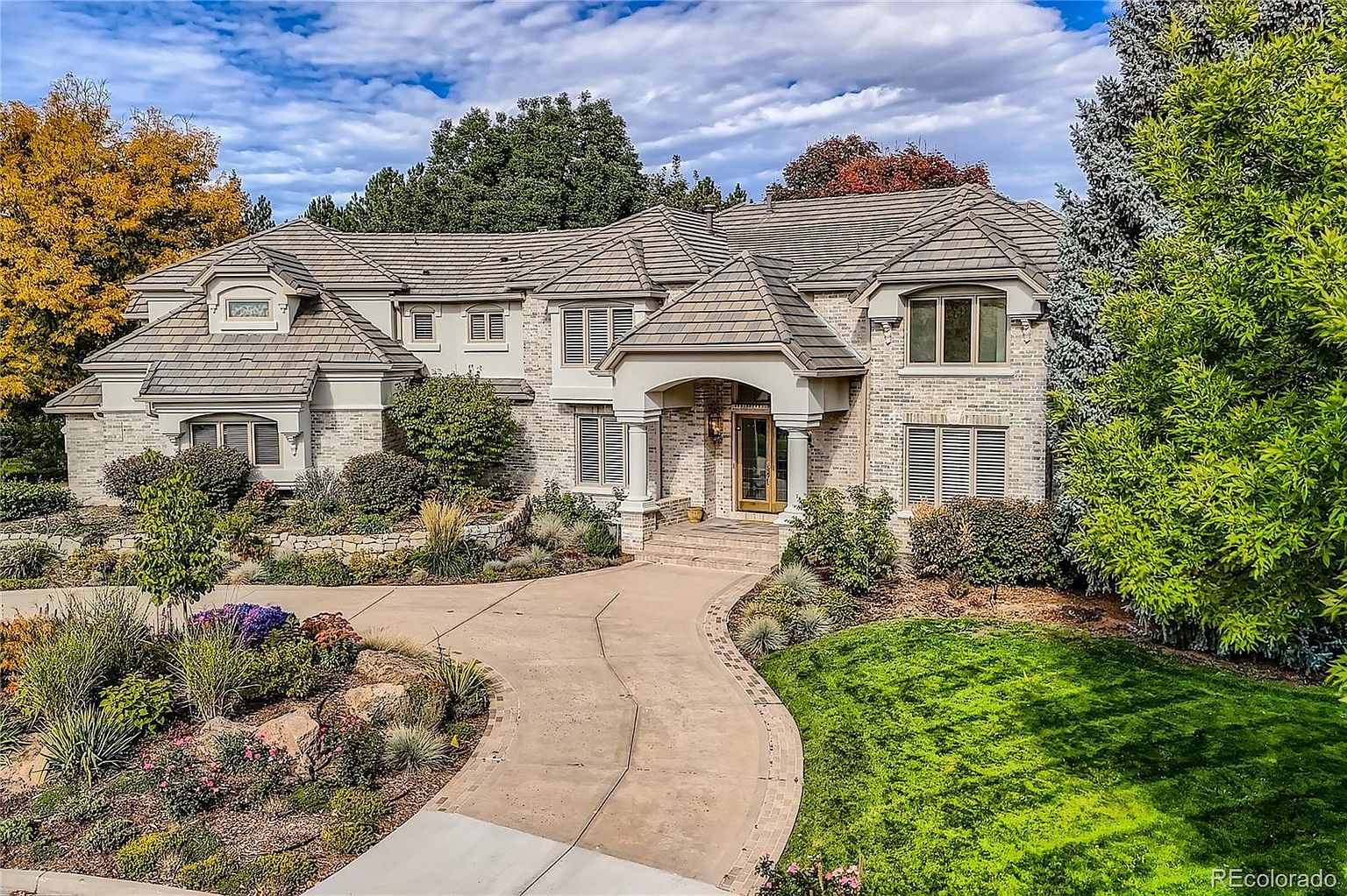 This elegant, multi-story brick estate features a sophisticated architectural design with a prominent arched entryway and a sweeping, paved driveway. The home is surrounded by lush, professionally landscaped gardens, mature trees, and a vibrant lawn, creating a welcoming and upscale curb appeal. The perspective is captured from a slightly elevated angle, showcasing the expansive facade and the harmonious blend of traditional brickwork with modern roof lines.
