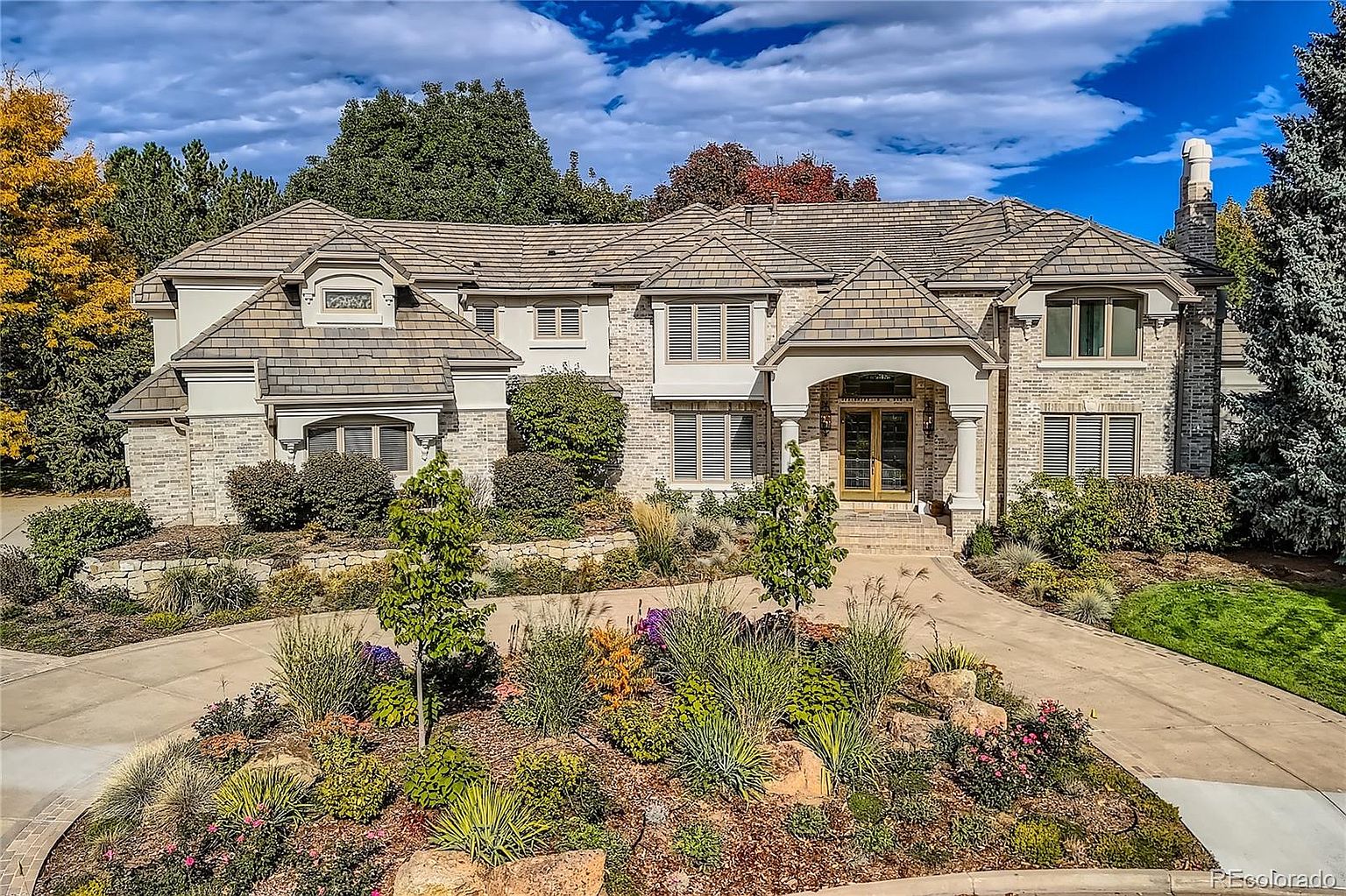 This grand, multi-story brick estate features a sophisticated architectural design with a prominent arched entryway and a sweeping concrete driveway. The front yard is meticulously landscaped with a variety of colorful perennials, ornamental grasses, and mature trees, creating a welcoming and upscale curb appeal. The perspective is a wide-angle, eye-level shot that captures the full breadth of the home's facade against a vibrant blue sky.