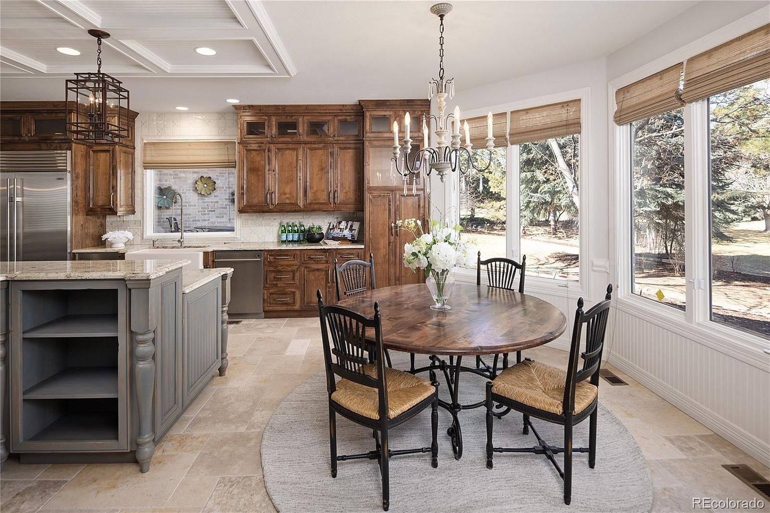 This elegant kitchen features a blend of rustic wood cabinetry and a contrasting grey-painted island, creating a warm and inviting atmosphere. A round wooden dining table sits on a circular rug under a classic chandelier, positioned perfectly to enjoy the natural light from the large windows. The space is well-appointed with high-end finishes, including stone tile flooring and a coffered ceiling, offering a sophisticated yet comfortable living environment.