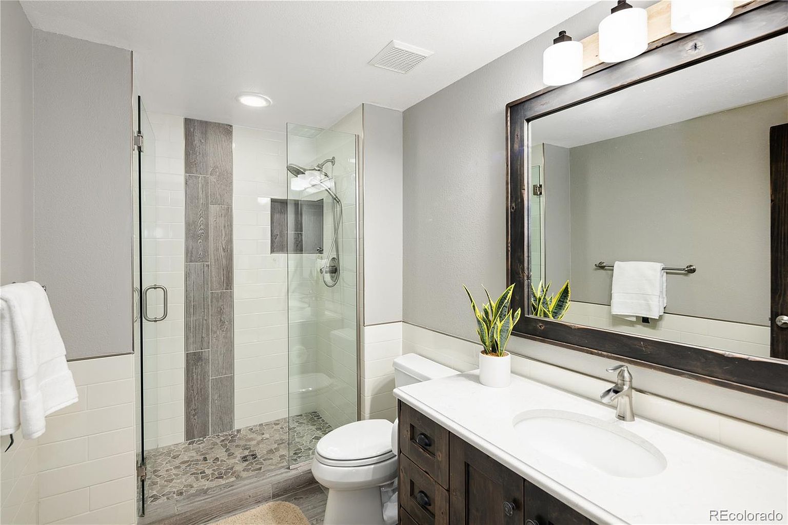 This modern bathroom features a sleek glass-enclosed walk-in shower with a pebble-stone floor and a decorative wood-look tile accent wall. The vanity is topped with a clean white countertop and paired with a rustic, dark-stained wood-framed mirror that adds a touch of warmth to the space. The overall aesthetic is a sophisticated blend of contemporary design and rustic charm, creating a spa-like atmosphere.