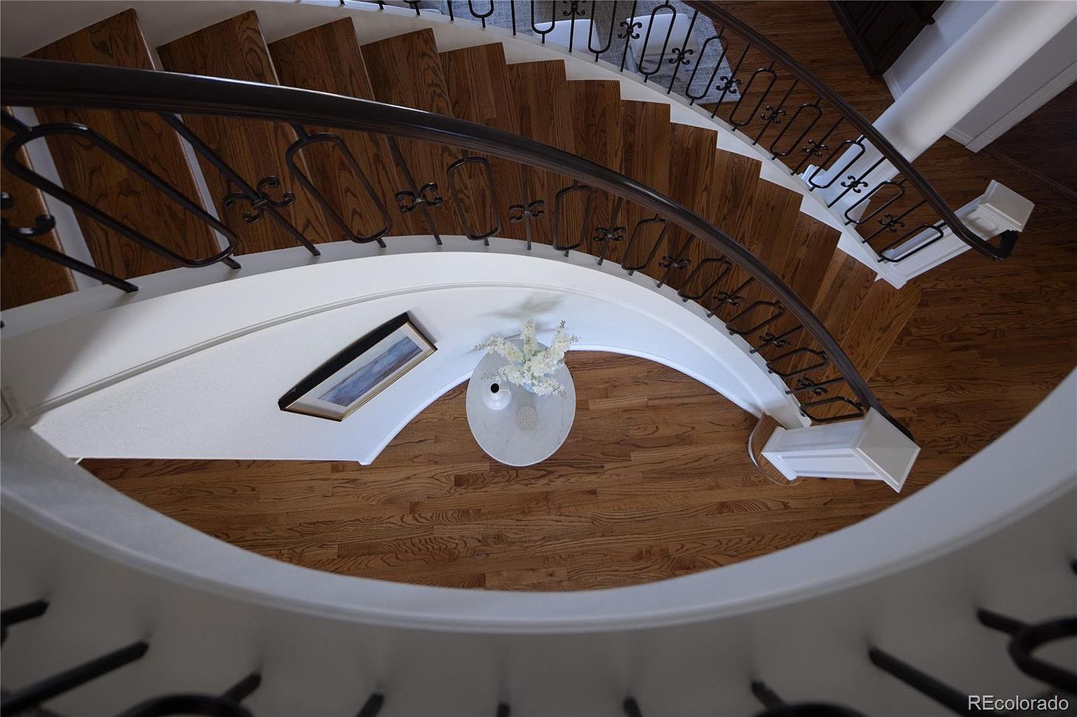 This high-angle, top-down perspective captures a graceful, curved staircase with rich hardwood treads and intricate wrought-iron balusters. Below, a small, elegant white pedestal table holds a delicate floral arrangement, complemented by a framed piece of art on the curved wall. The composition emphasizes the architectural flow and sophisticated design of the home's entryway.