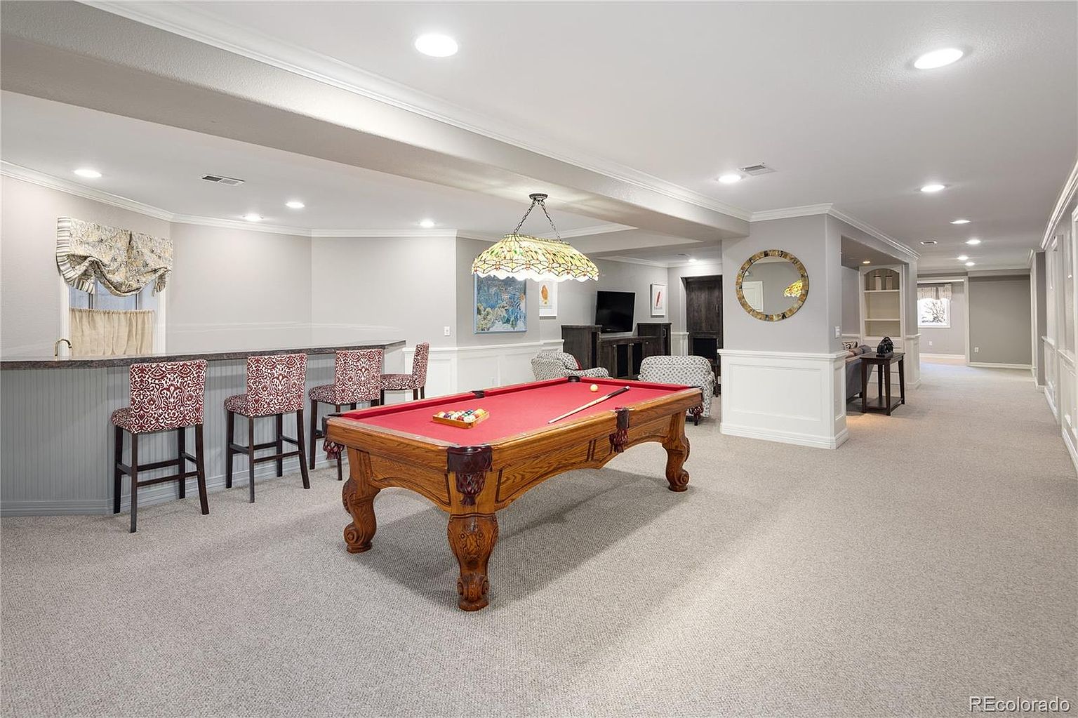 This spacious finished basement features a classic game room layout with a prominent red-felt pool table as the centerpiece. The area includes a built-in bar with patterned barstools, recessed lighting, and a neutral color palette that creates a comfortable, inviting atmosphere for entertainment. The open floor plan extends into a secondary seating area, showcasing a versatile space perfect for hosting guests.