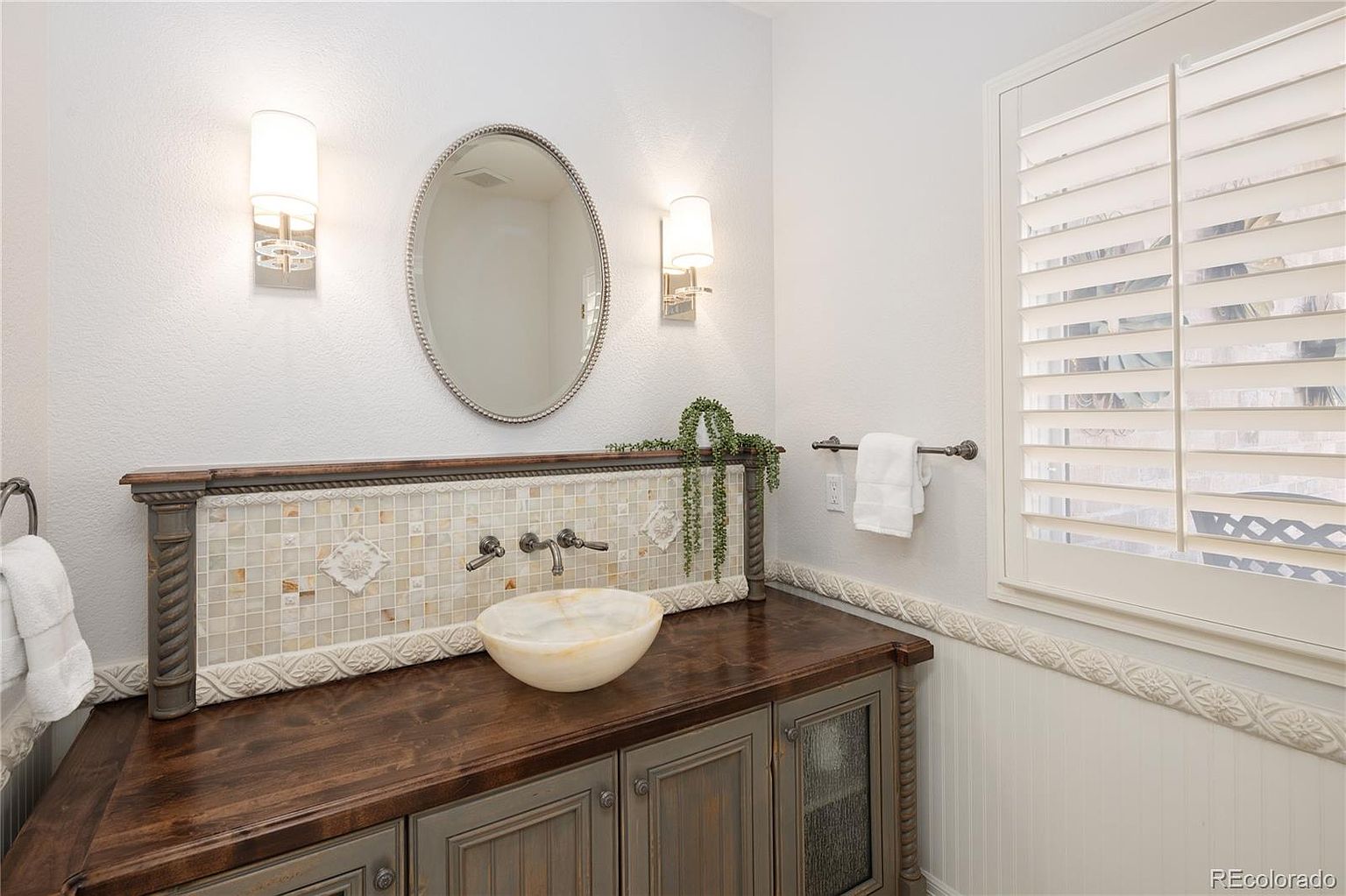This elegant powder room features a unique stone vessel sink set atop a rustic, dark-stained wooden vanity with decorative cabinetry. The wall is accented with a beautiful mosaic tile backsplash, a classic oval mirror, and matching wall sconces, all framed by white wainscoting and plantation shutters. The overall aesthetic is a sophisticated blend of farmhouse charm and refined traditional design.