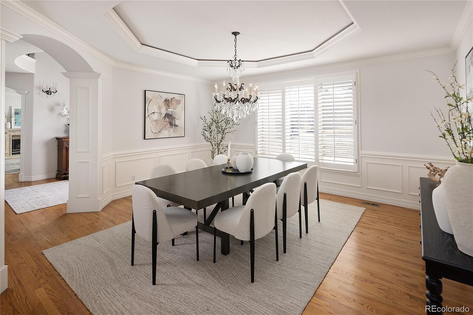 This elegant dining room features a dark wood table surrounded by six modern, textured white chairs, all set atop a neutral area rug. The space is defined by sophisticated wainscoting, a dramatic crystal chandelier hanging from a tray ceiling, and plantation shutters that allow soft natural light to filter in. The overall aesthetic is refined and polished, creating a welcoming atmosphere for formal gatherings.