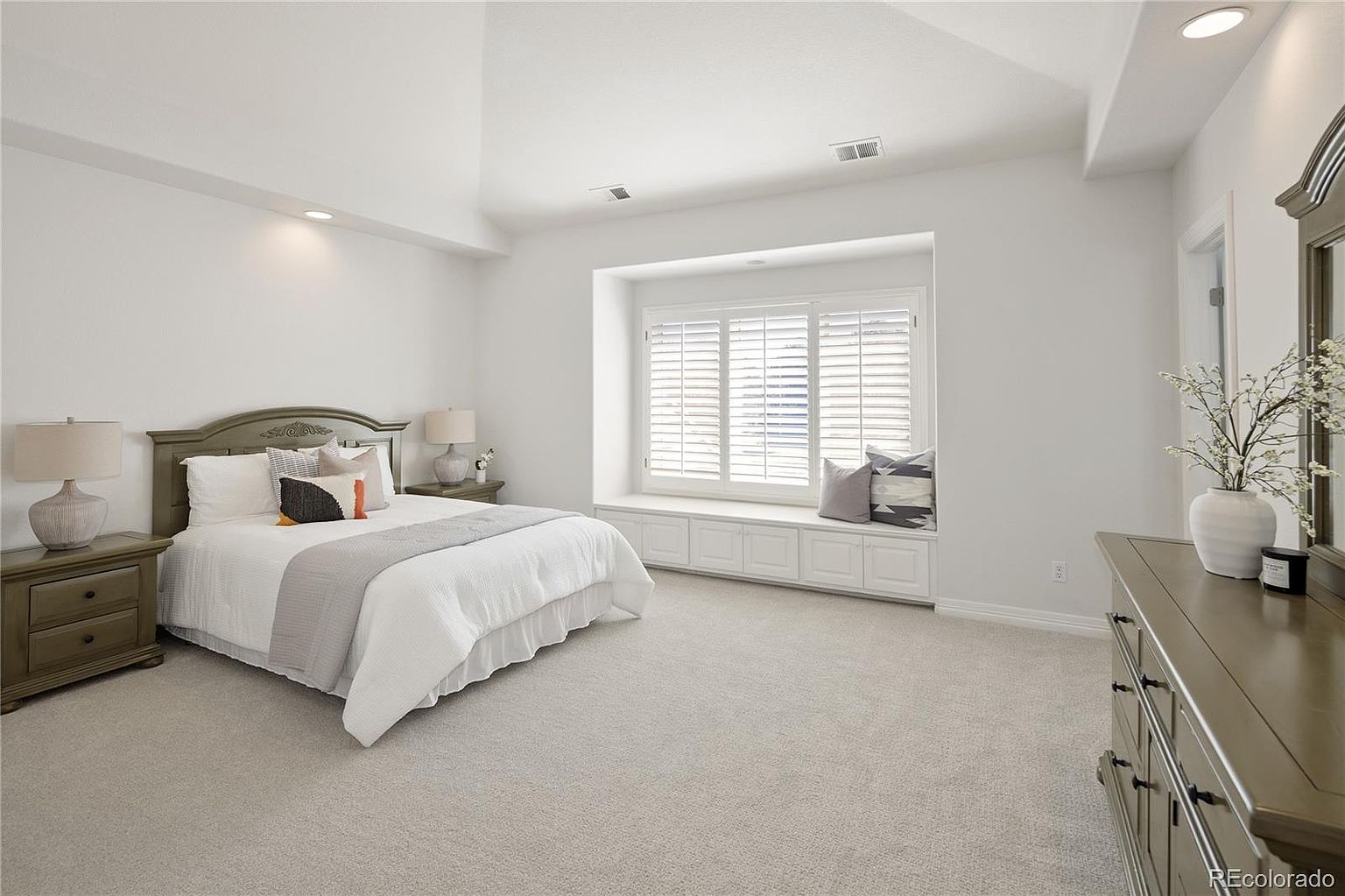 This spacious primary bedroom features a high vaulted ceiling, neutral wall tones, and plush carpeting, creating a serene and airy atmosphere. A prominent bay window with built-in bench seating serves as a focal point, complemented by a classic bed frame and matching nightstands. The room is well-lit with recessed lighting and offers a clean, sophisticated aesthetic suitable for a modern home.