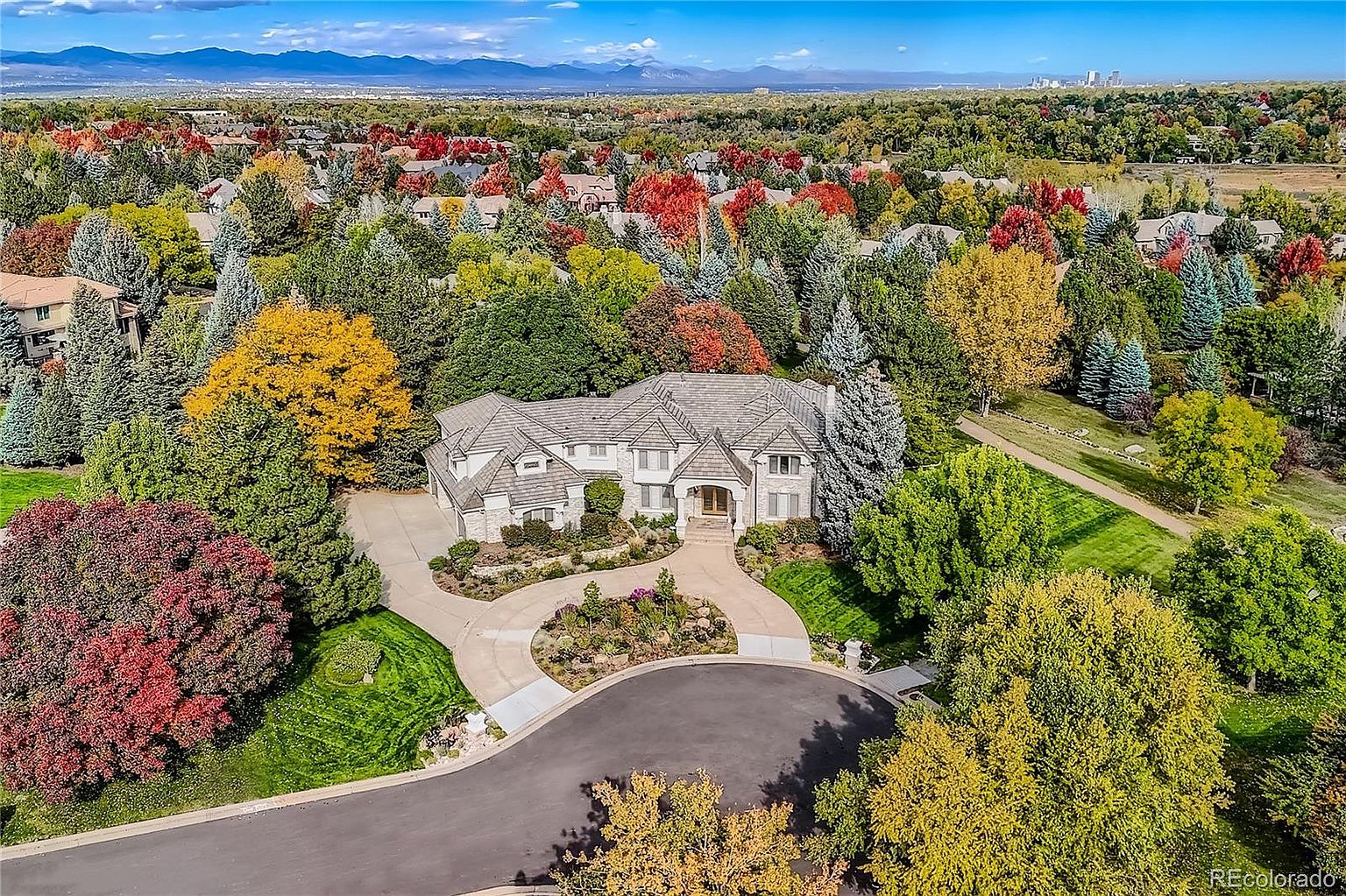 This high-angle aerial shot captures a sprawling, luxury suburban estate nestled within a lush, tree-filled landscape during autumn. The home features a classic architectural design with a multi-gabled roof, a circular driveway, and a well-manicured front garden, all set against a backdrop of distant mountains and a clear blue sky. The vibrant fall foliage surrounding the property enhances its curb appeal and serene, upscale atmosphere.