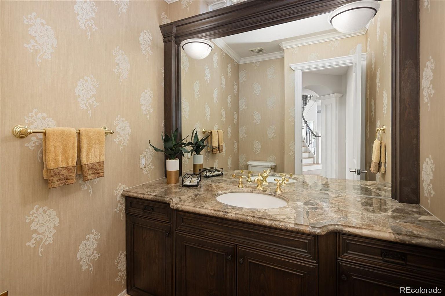 This elegant powder room features a rich, dark wood vanity topped with a luxurious brown marble countertop and an undermount sink. The walls are adorned with a sophisticated floral-patterned wallpaper, complemented by a large, framed mirror and classic gold-toned fixtures. The space exudes a traditional, refined aesthetic, offering a glimpse into the adjacent hallway and staircase.