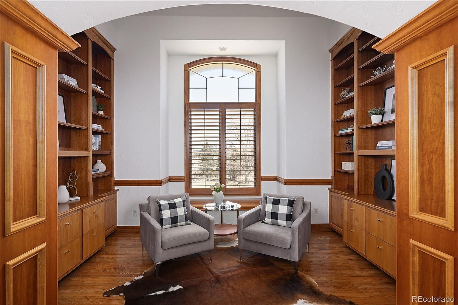 This elegant home office or reading nook features custom built-in wooden bookshelves flanking a large, arched window with plantation shutters. Two plush grey armchairs with buffalo check pillows are centered on a cowhide rug, creating a cozy and sophisticated seating area. The warm wood tones of the cabinetry and flooring, combined with the architectural archway, provide a refined and inviting atmosphere.