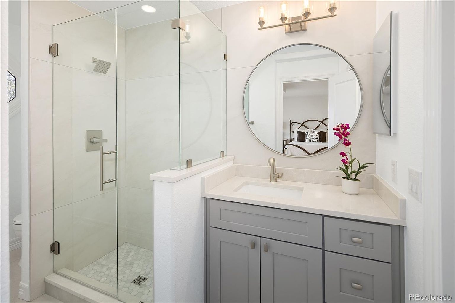This modern bathroom features a sleek gray vanity with a white stone countertop, a circular mirror, and a glass-enclosed walk-in shower with light-colored tile. The space is bright and clean, with a minimalist aesthetic that highlights the contemporary fixtures and neutral color palette. The perspective captures the vanity and shower area, reflecting a glimpse of the adjacent bedroom in the mirror.