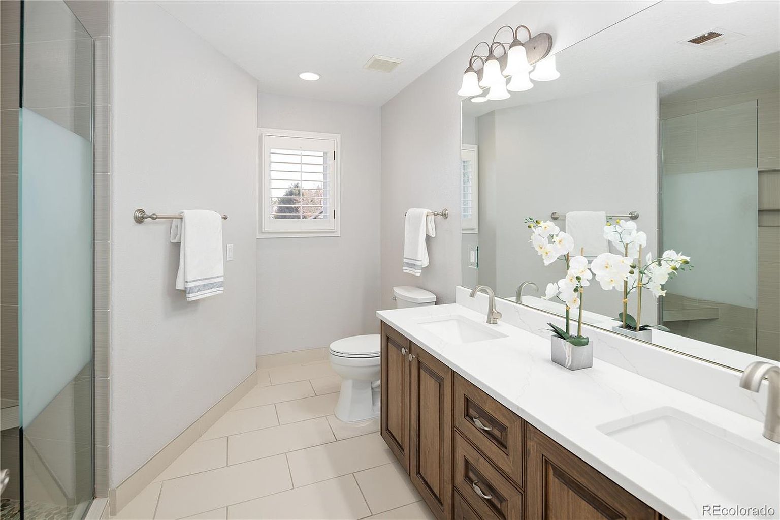 This bright and modern primary bathroom features a double vanity with dark wood cabinetry, white quartz countertops, and dual undermount sinks. A large, frameless mirror spans the wall above the vanity, complemented by a stylish multi-light fixture and a glass-enclosed shower. The space is finished with light-colored floor tiles and a window with plantation shutters, creating a clean, airy, and sophisticated atmosphere.