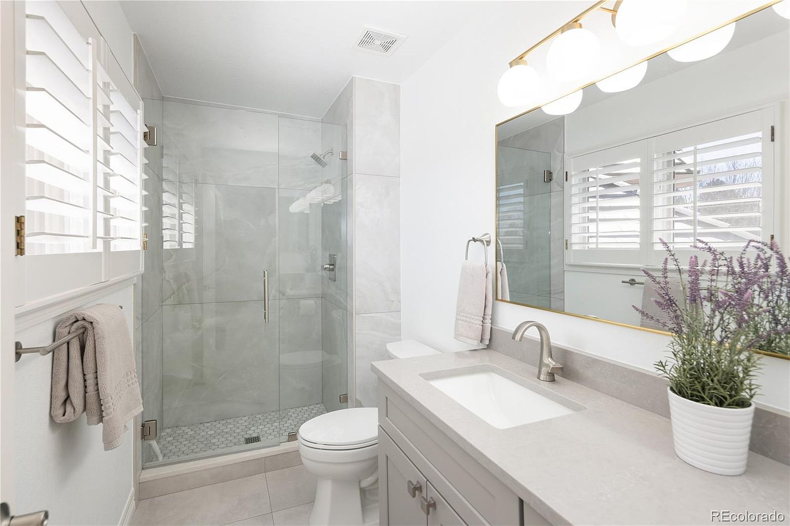 This bright and modern bathroom features a sleek vanity with a quartz countertop, a glass-enclosed walk-in shower with light-toned tile, and a crisp white toilet. The space is accented by a gold-framed mirror, contemporary globe lighting, and plantation shutters that allow for natural light. A potted lavender plant adds a touch of color to the clean, neutral aesthetic, creating a fresh and inviting atmosphere.